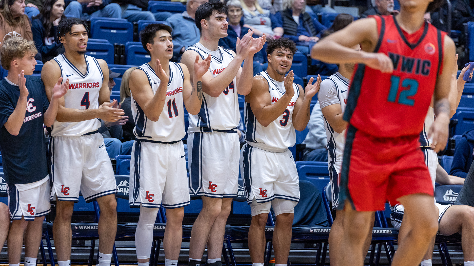 LC State men’s basketball players in white Warriors uniforms stand and applaud from the bench during a game, while an opposing player in a red uniform runs past in the foreground and spectators watch from the seats behind them.