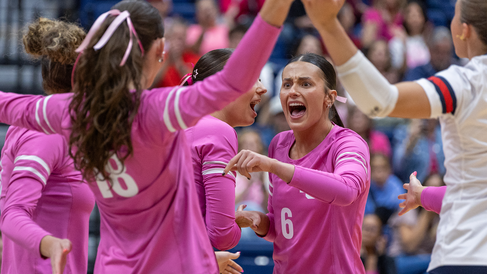 LC State Volleyball players in pink jerseys celebrate together on the court during Pink Night, with pink ribbons in their hair and fans dressed in pink visible in the background.