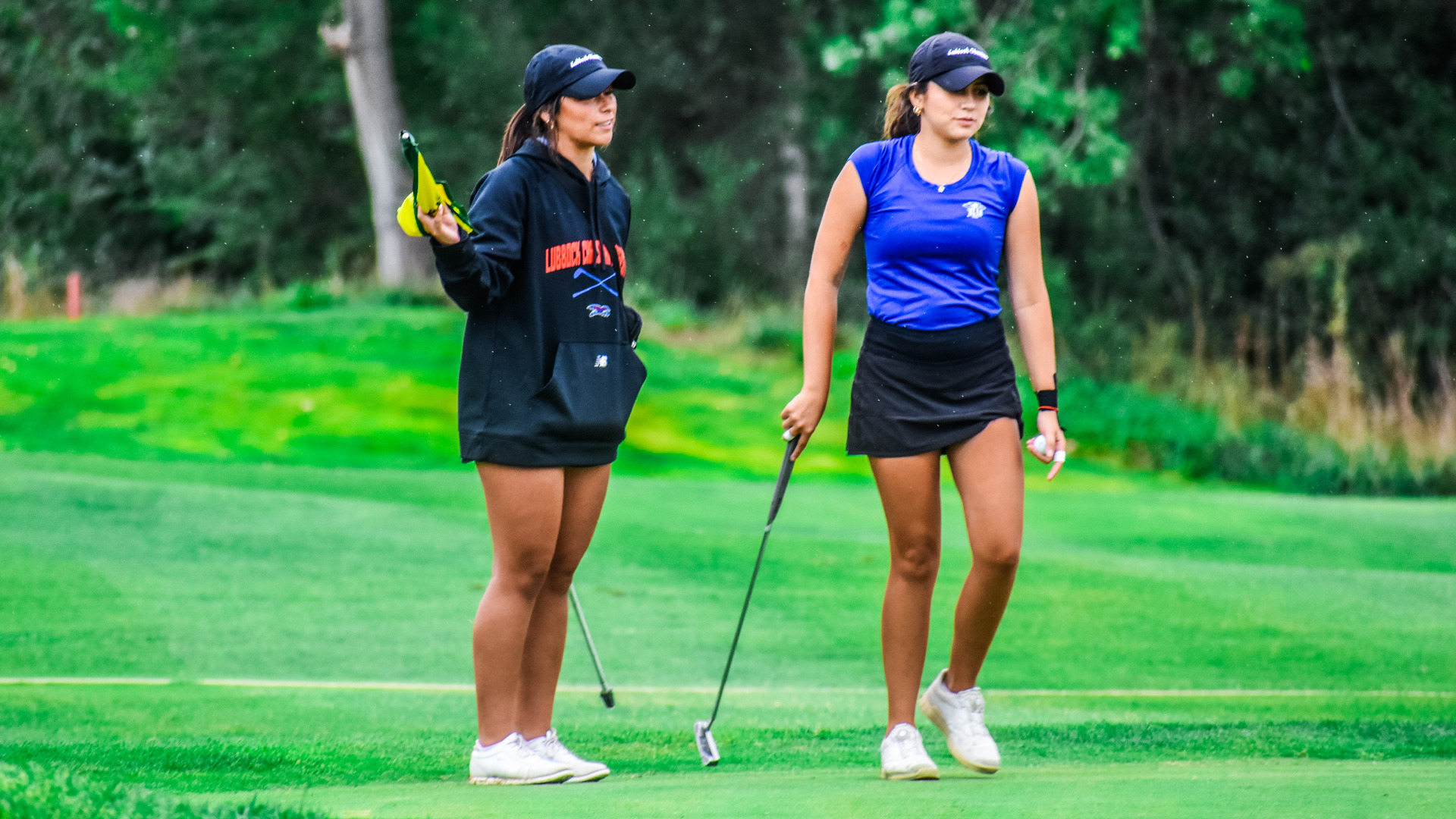 Two female golfers standing on green fairway, one in black jacket holding yellow flag pin and the other in blue sleeveless top and black skirt holding putter, with wooded area in background