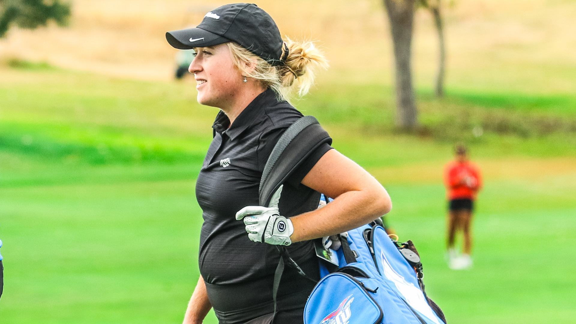 Female golfer in black Nike cap and black sleeveless polo carrying blue and white golf bag with clubs on green fairway course, smiling while looking off to the side with another golfer visible in blurred background