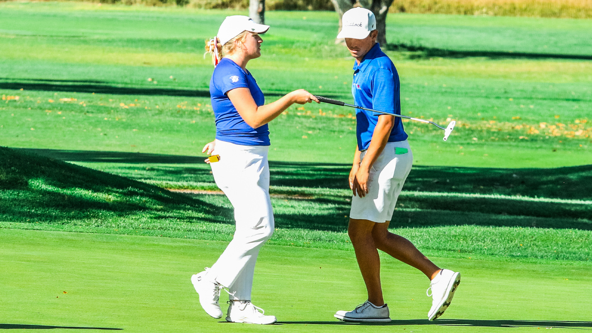 A female golfer and male head coach in matching blue team polos and white caps walking together on green fairway, one in white pants and one in white skirt, with player on left holding putter and appearing to discuss strategy with teammate