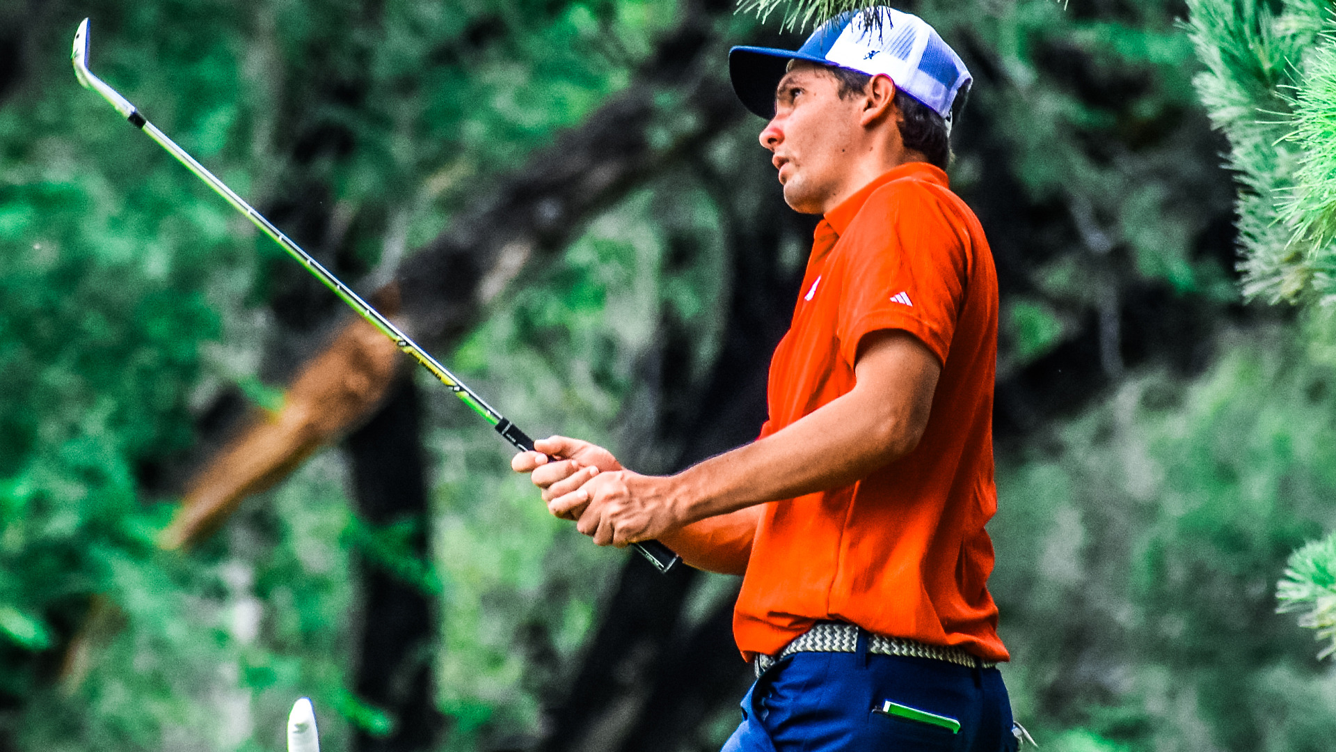Male golfer in orange shirt and blue-white cap watching shot after swing, holding club in follow-through position with green trees and foliage in blurred background