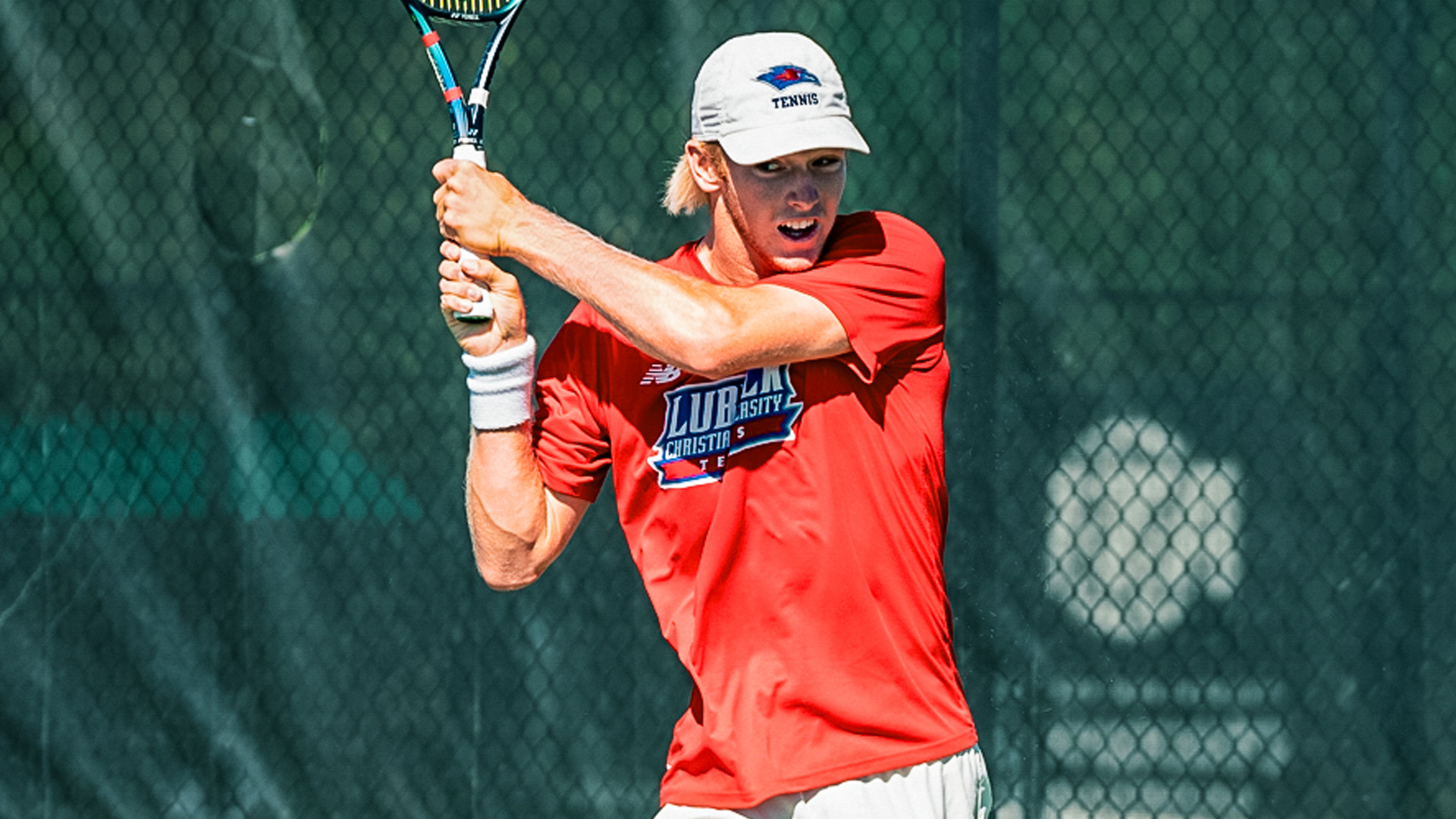 Male tennis player in red Lubbock Christian shirt and white cap executing two-handed backhand shot on outdoor court with green chain-link fence in background