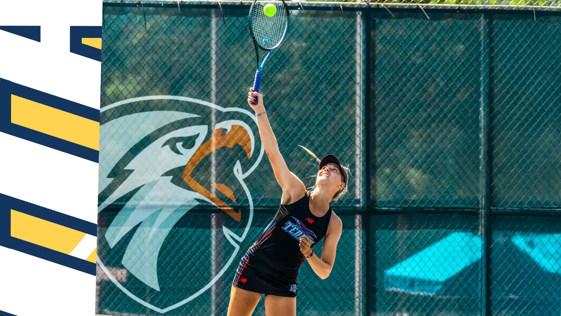 Female tennis player in black uniform serving the ball on outdoor court with green fence backdrop featuring team logo, reaching up with racket to make contact with yellow tennis ball