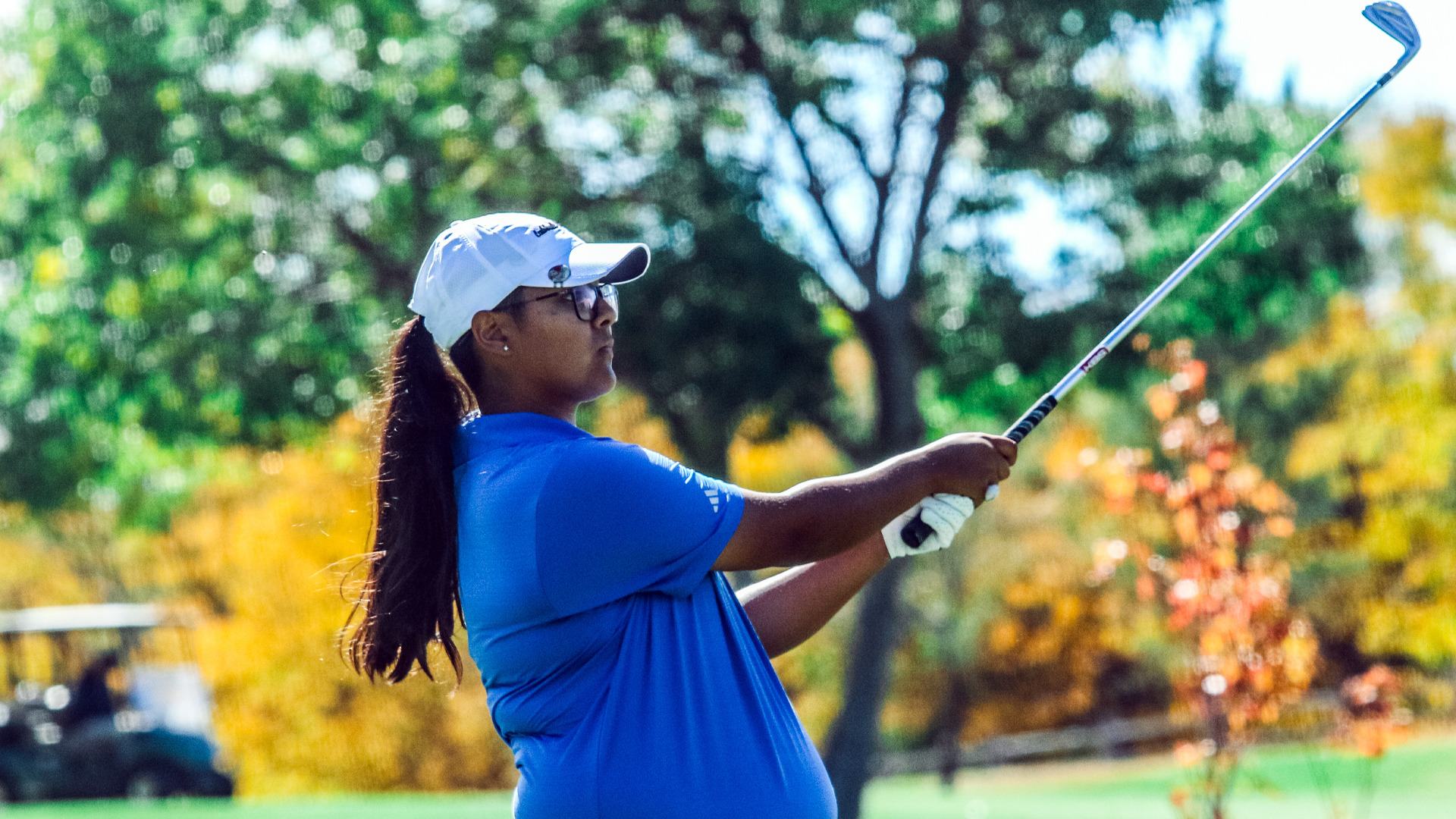 Female golfer in blue shirt, white cap and glasses completing follow-through of golf swing on course with vibrant green, yellow and orange autumn foliage and trees in soft focus background