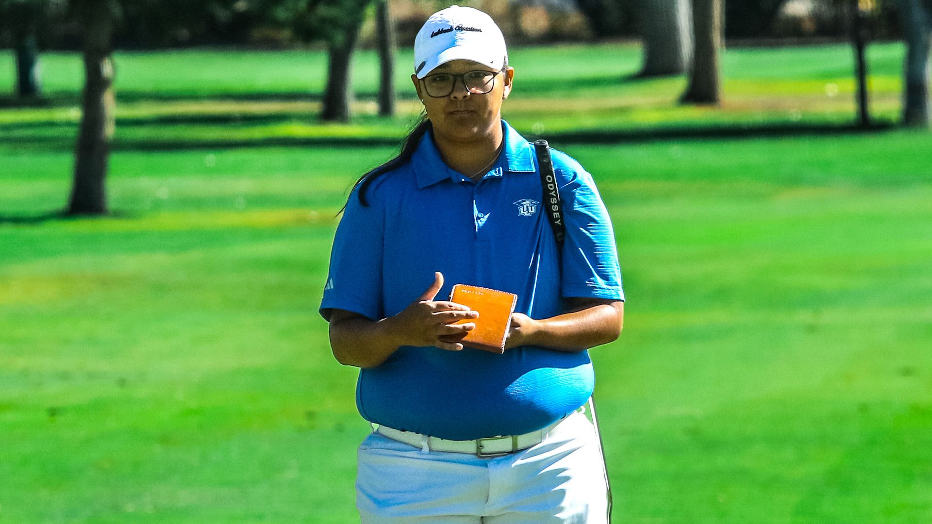 Female golfer in blue Lubbock Christian polo shirt, white cap and sunglasses holding orange yardage book while standing on green fairway with trees in soft focus background