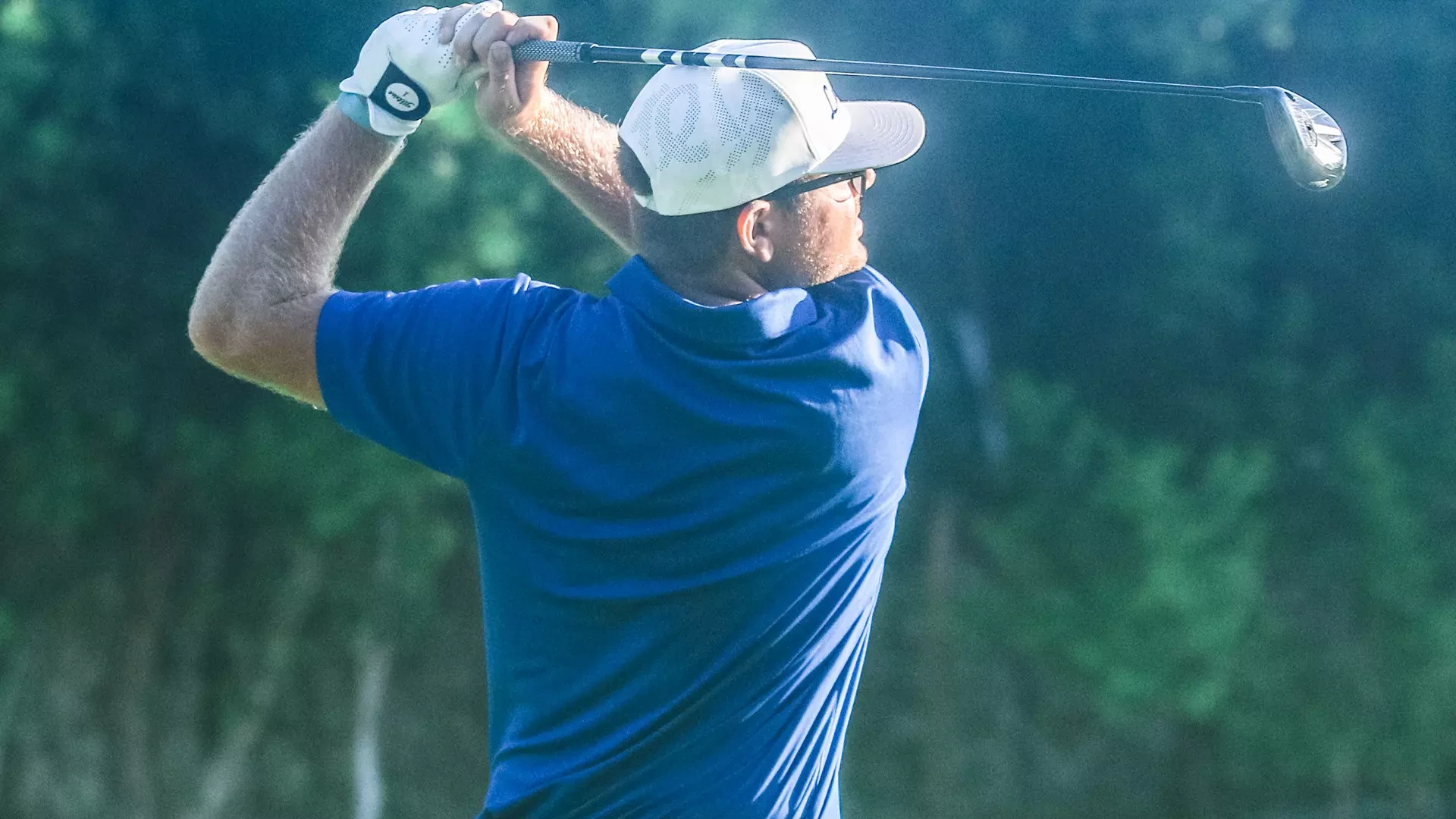 Golfer in blue shirt and white cap completing follow-through of golf swing against blurred green background
