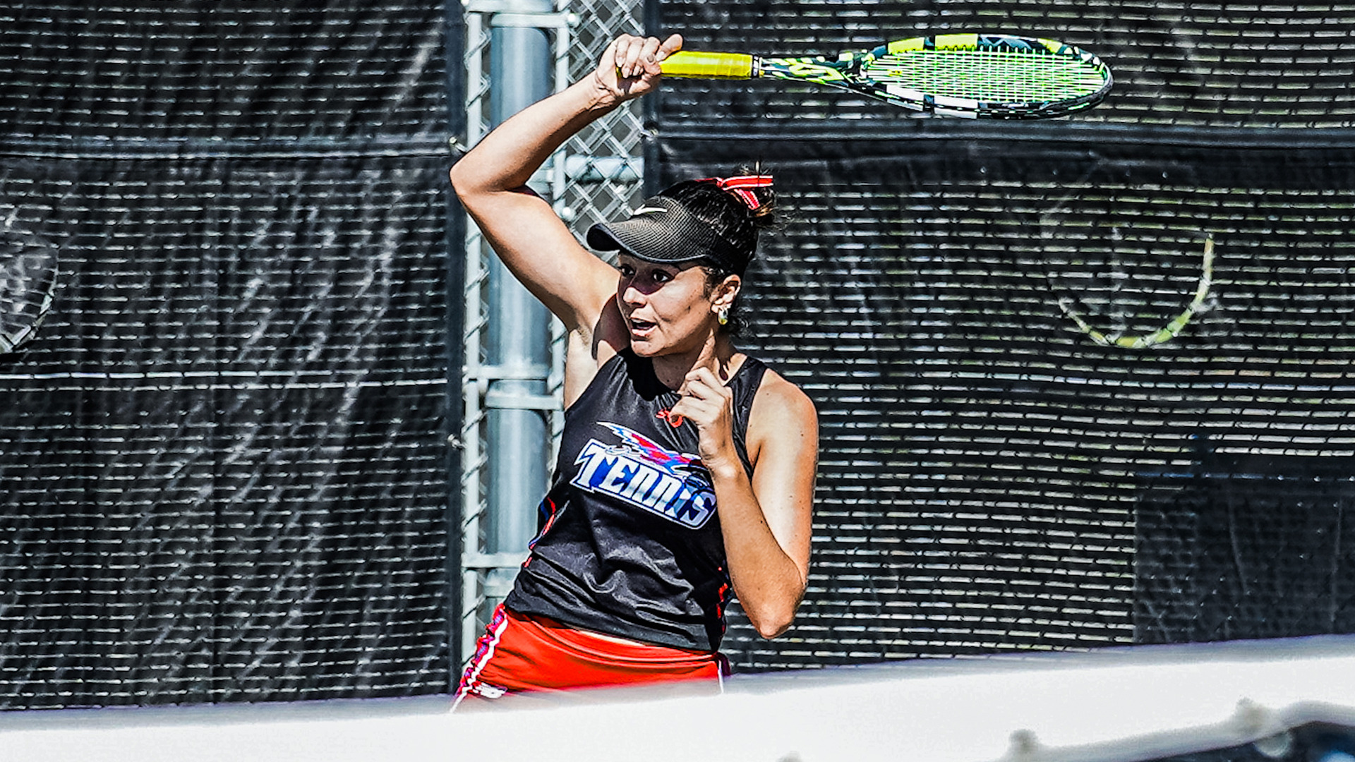 Tennis player in black and red uniform with visor preparing to serve, raising yellow and green racket overhead at outdoor court