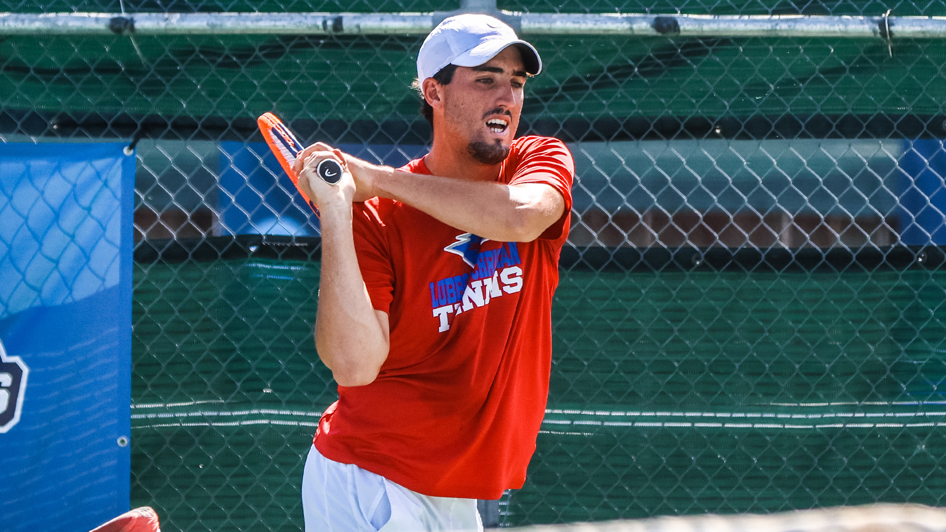 Tennis player in red shirt and white cap hitting forehand return during match at outdoor court with chain-link fence in background