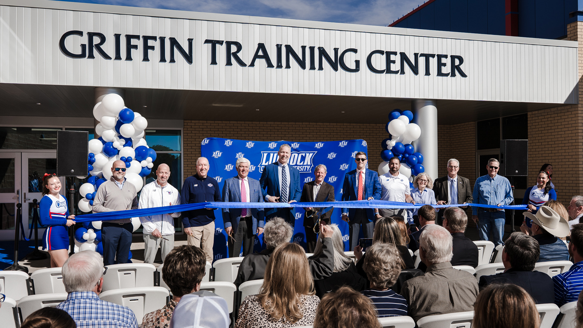 Ribbon cutting ceremony at Griffin Training Center with officials and attendees holding large blue ribbon in front of building entrance decorated with blue and white balloon displays