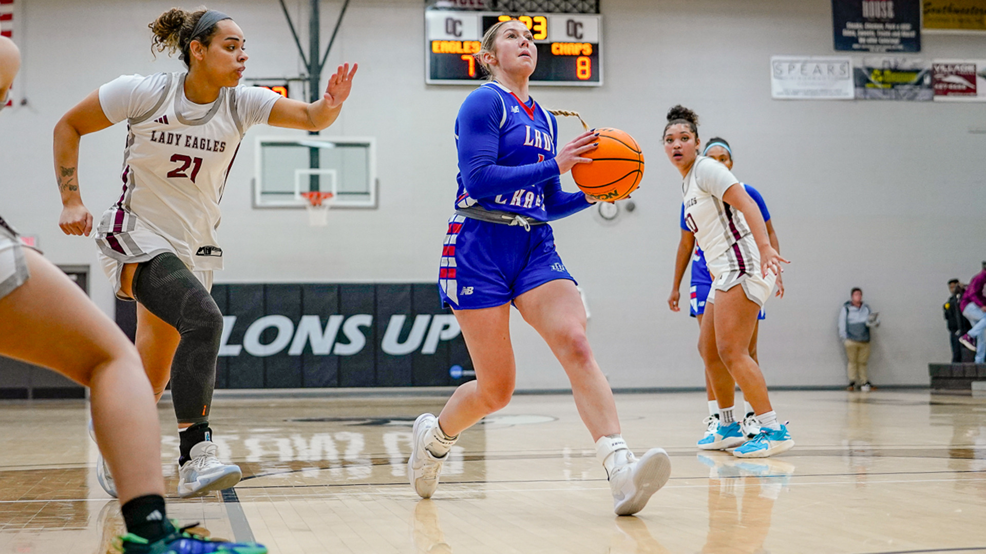 Action photograph from a Lubbock Christian University Lady Chaps basketball game showing a player in a blue LCU uniform driving toward the basket while holding a Wilson basketball. The player is being defended by opponents in white 