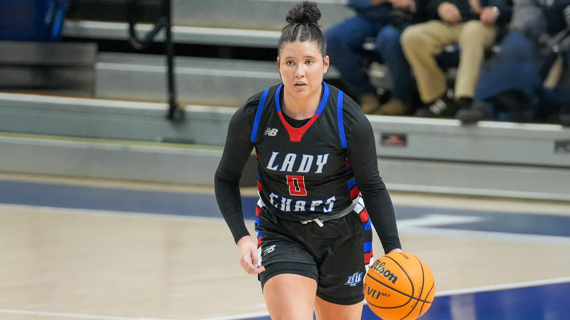 Action photograph from a Lubbock Christian University Lady Chaps basketball game showing a player in a black Lady Chaps uniform number 0 with blue and red trim dribbling a Wilson basketball down the court with a focused expression. Spectators are visible in the blurred background seating area. The photograph captures a moment during competitive play.