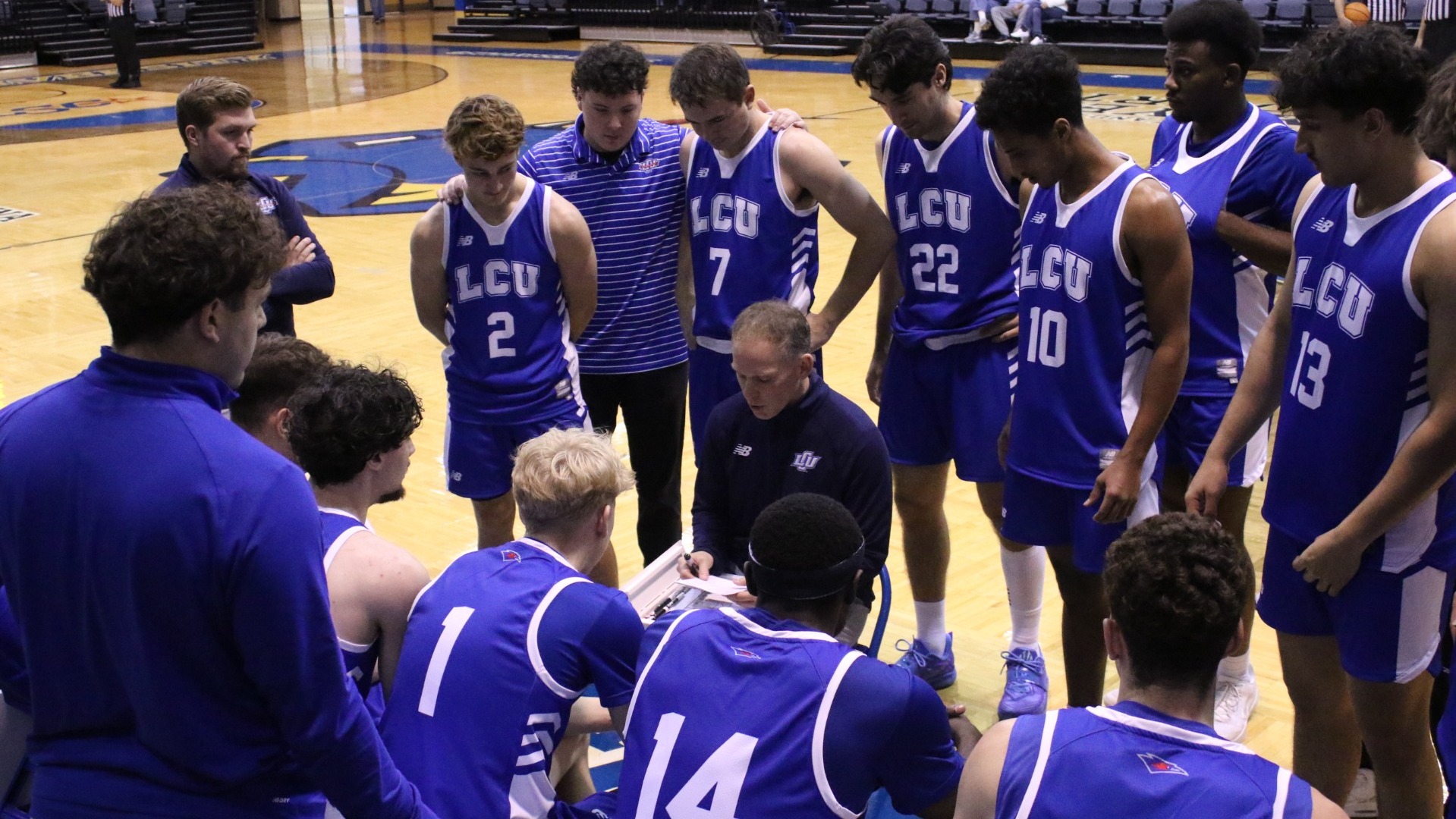 This is a photograph of Lubbock Christian men's basketball coach Jason Imes (navy blue shirt, center) talking to the Chaparral team (wearing blue jerseys) during a timeout at Bill Greehey Arena.