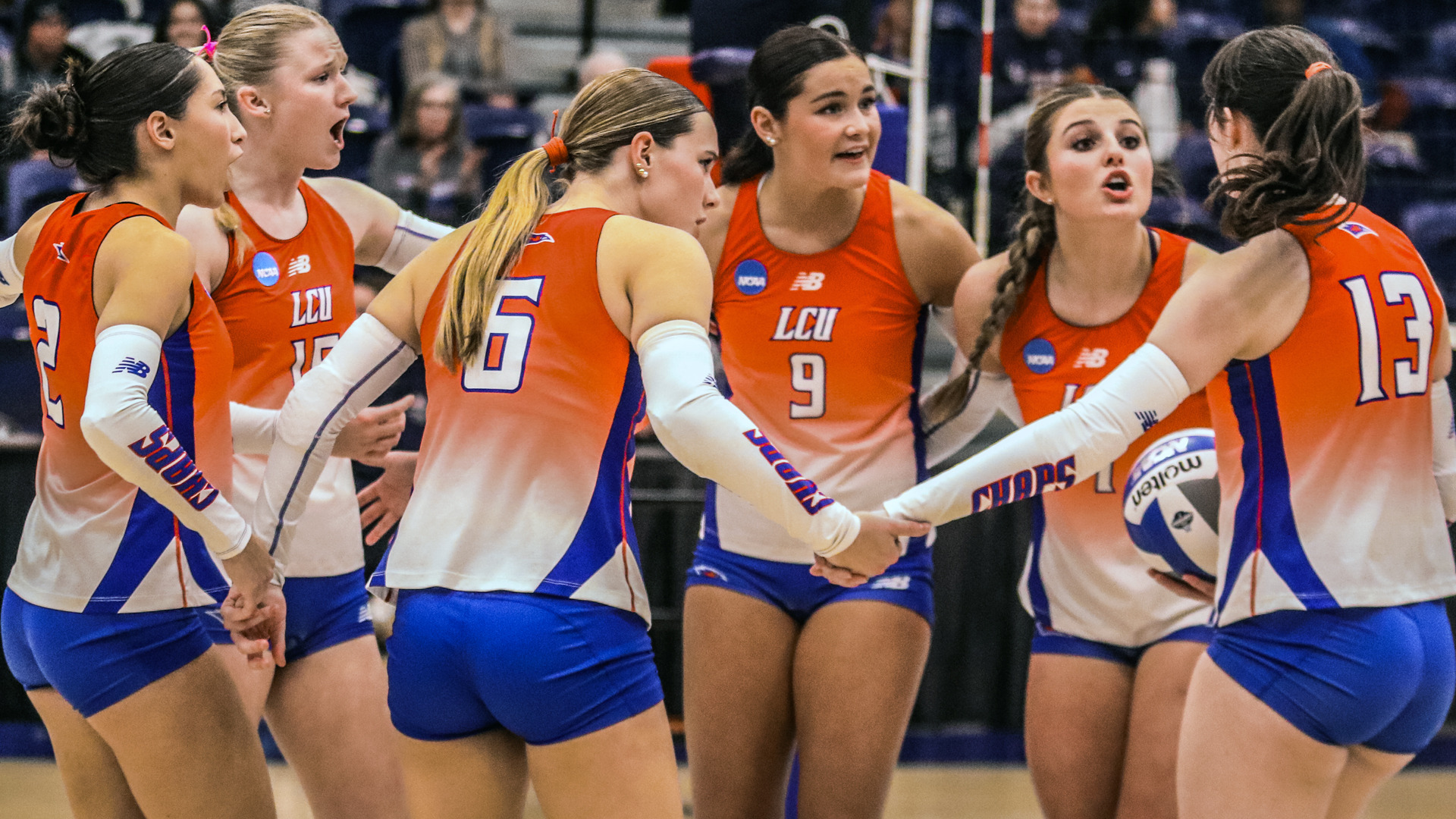Action photograph from a Lubbock Christian University Lady Chaps volleyball match showing multiple female volleyball players in orange and white gradient LCU uniforms with blue shorts and white arm sleeves celebrating together on the court during NCAA tournament play. The players wear various jersey numbers including 2, 10, 5, 9, and 13, with NCAA patches and New Balance logos visible on their uniforms. The team displays excited expressions and engaged body language, creating an energetic moment of celebration. Spectators fill the background bleachers, watching the game intently. The photograph captures the team's unified enthusiasm during competitive play.