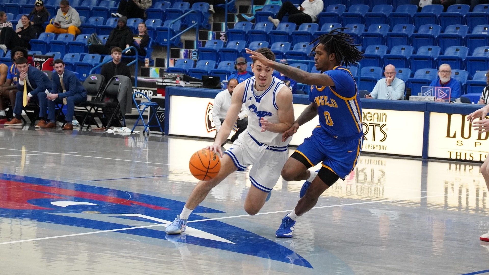 This is a photograph of Lubbock Christian forward Amondo Miller Jr. (white jersey, #8) dribbling a basketball past an Angelo State defender (blue jersey, #0) in a game at the Rip Griffin Center. 