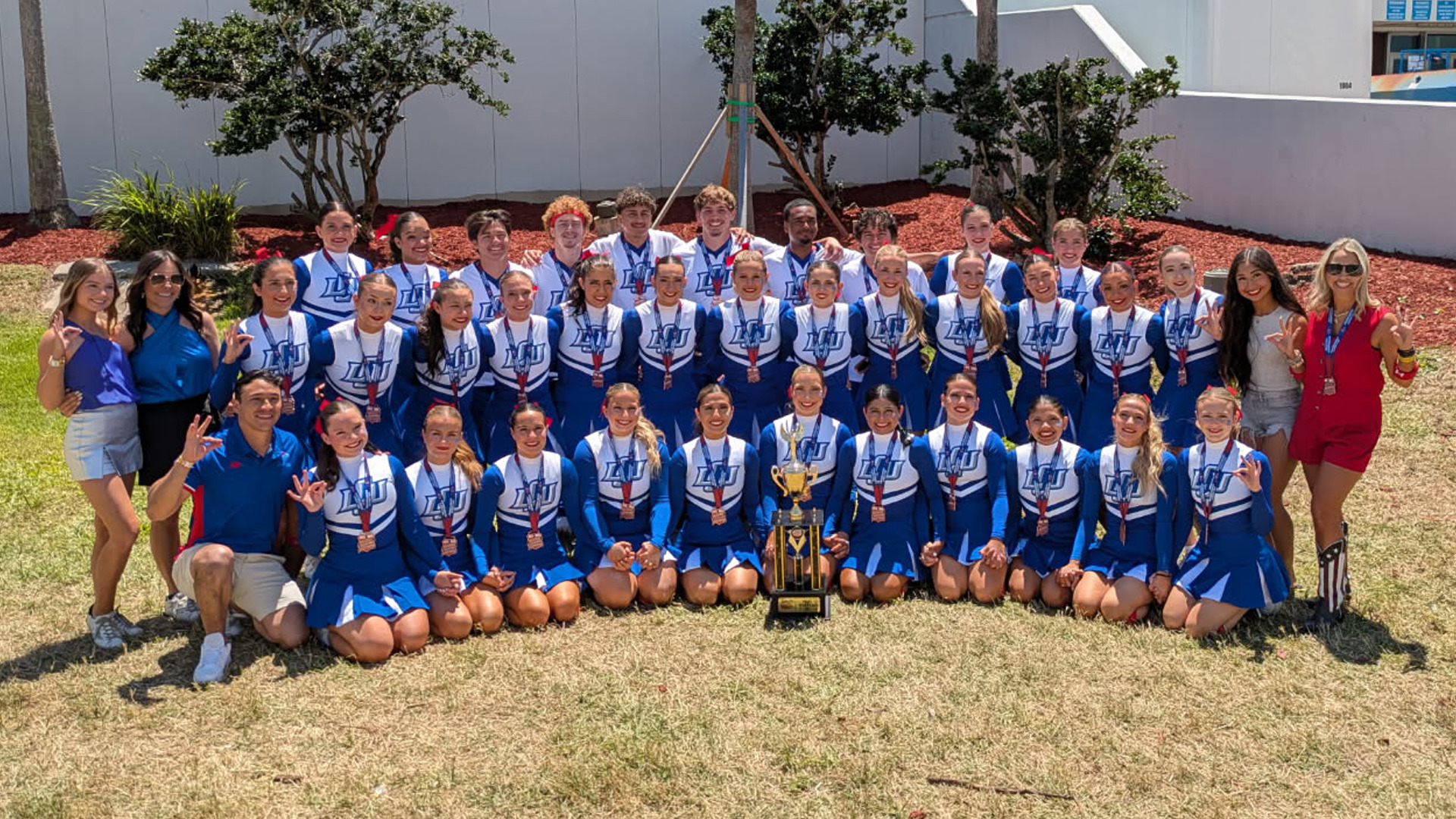 A large cheerleading team from Lubbock Christian University poses outside in front of a white building with shrubs and red mulch. The team consists of male and female cheerleaders in blue and white LCU uniforms, standing and kneeling in rows. Most team members are holding up the “L” hand sign for LCU. Coaches and supporters in casual and team apparel stand on either side of the group. A large championship trophy is displayed prominently in front of the center row, indicating a significant achievement at a national competition. It is a sunny day, and the group appears celebratory and proud.