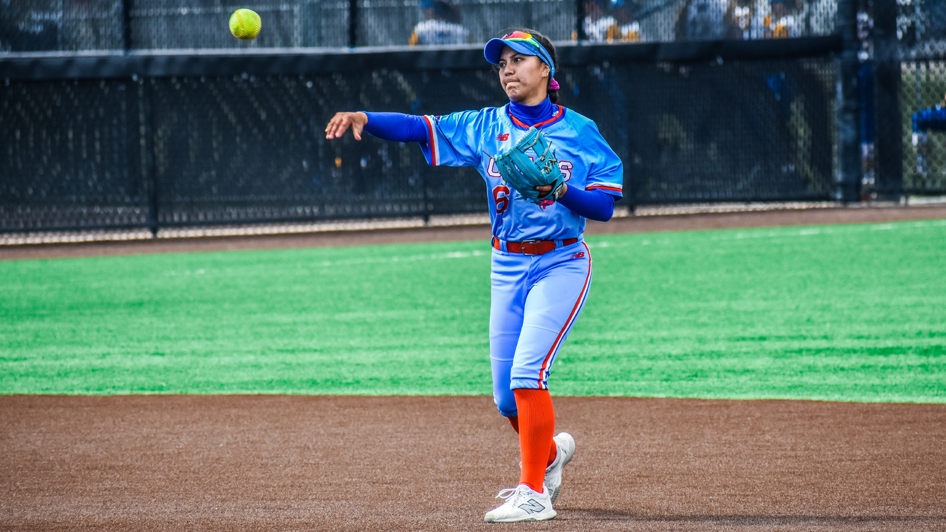 Lubbock Christian University softball player Kawehi Ili is pictured mid-action during a game. She wears a sky-blue uniform with red accents, featuring the number 6 on her jersey. Ili is on a turf field, positioned on the dirt portion near the infield, and is throwing a softball with her right hand while wearing a teal glove on her left. Her expression is focused as she completes the throw. She also wears a blue visor with reflective sunglasses and long blue sleeves under her jersey. The background includes a black chain-link fence and blurred spectators