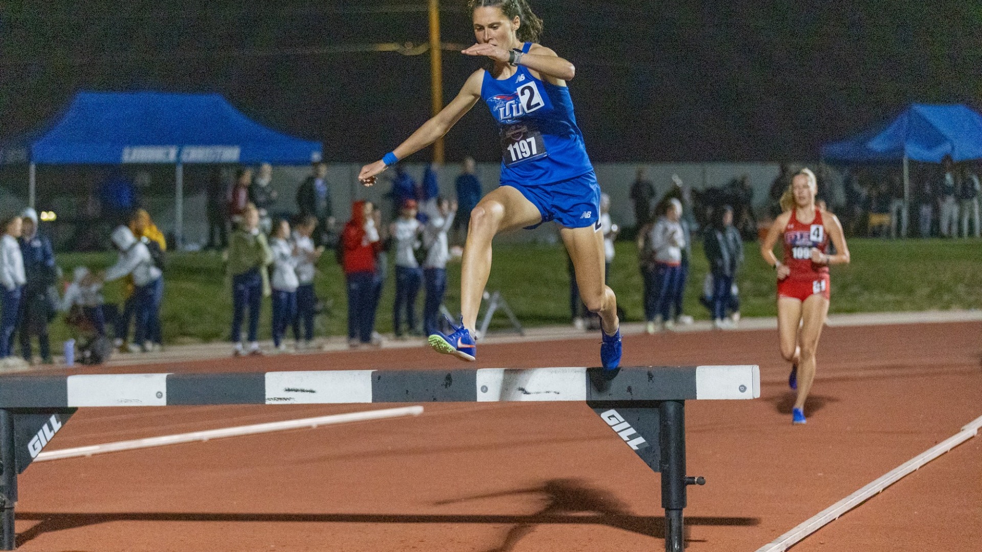 Action shot of Lubbock Christian University track athlete, Olivia Weider, clearing an obstacle in the 3000-meter steeplechase at the Lone Star Conference Outdoor Championships. She is seen in midair with her legs extended, as she is looking down ready to land. She is wearing a blue LCU track uniform