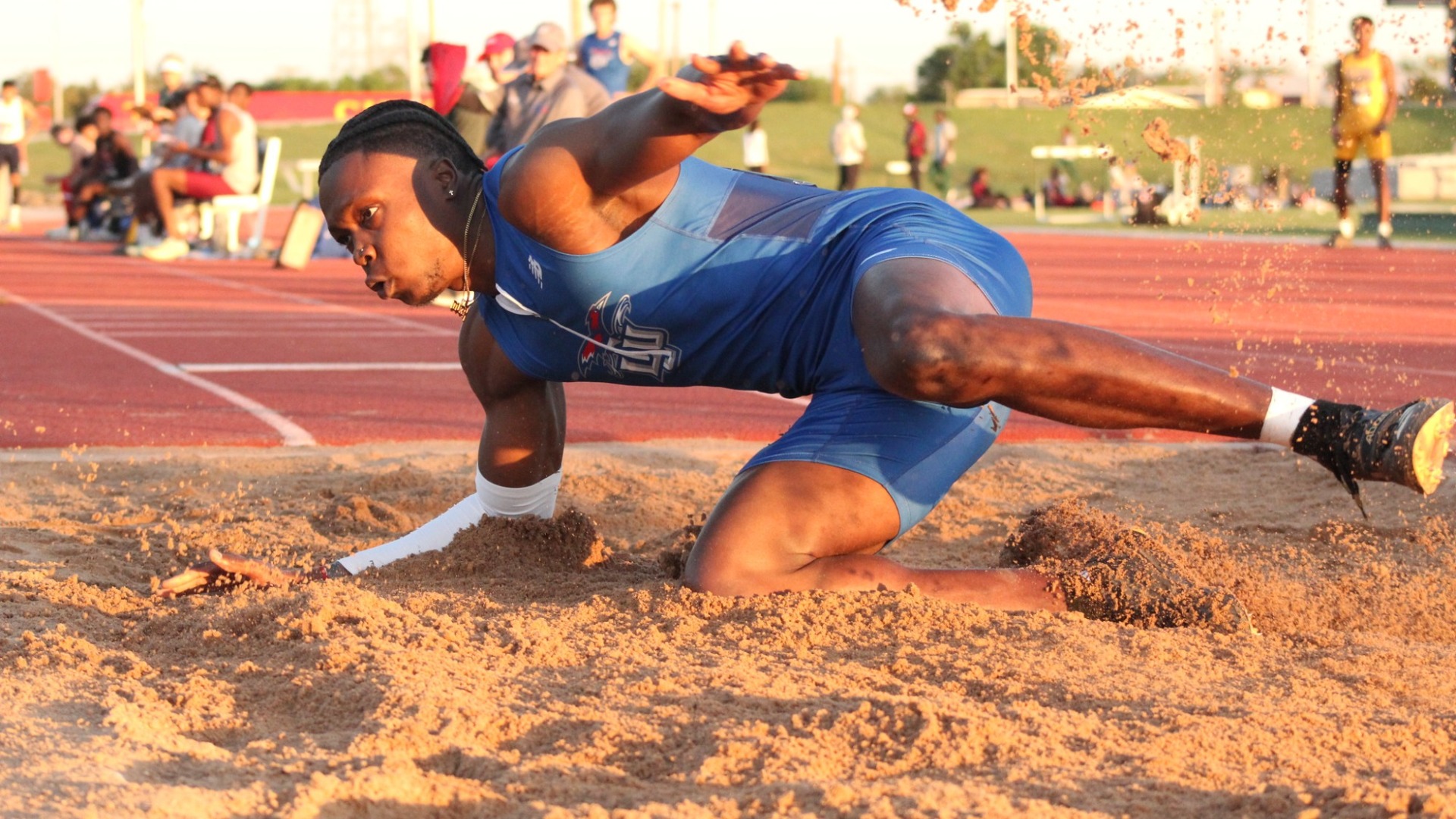 Action shot of Lubbock Christian University jumper, Kevin Washington, as he is seen laying in the sand following a long jump attempt. Washington is laying horizontal and is partially obscured by the sand. He is wearing a blue LCU tracksuit.