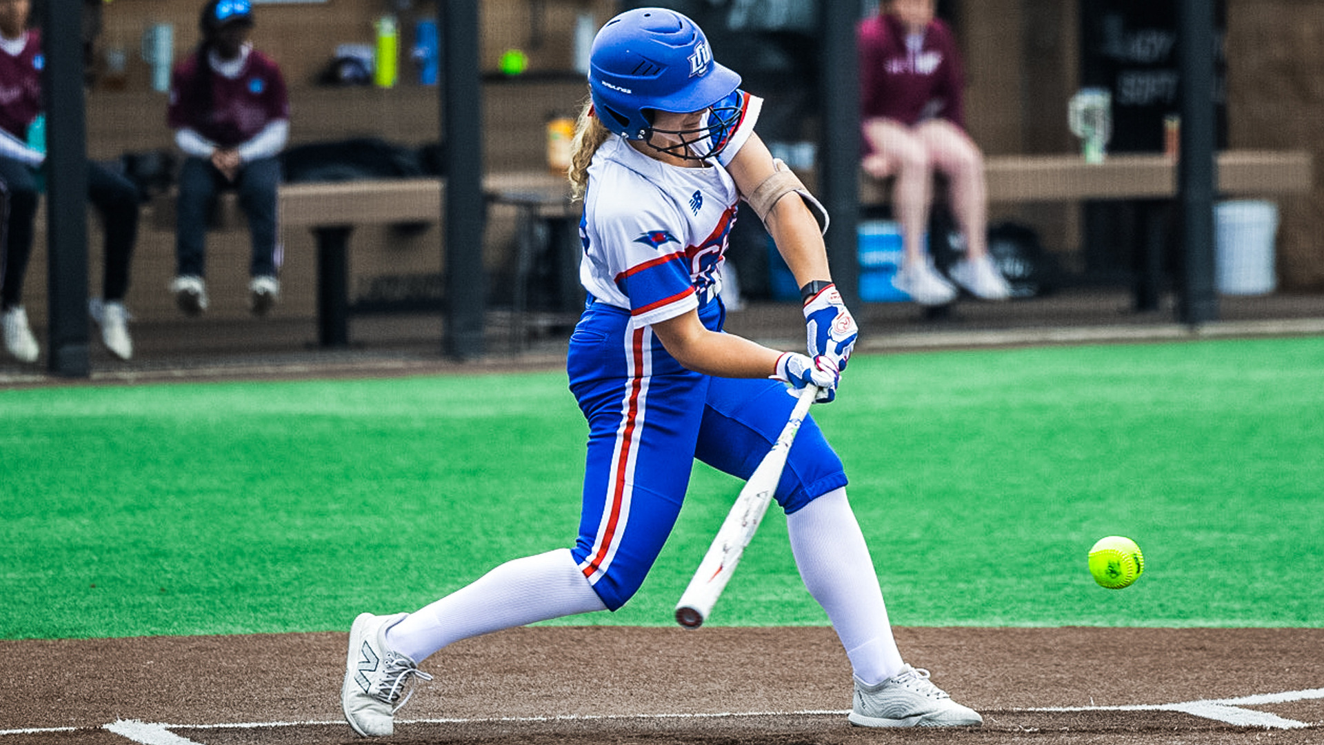 Lubbock Christian University softball player Kristen Boyd, wearing a blue helmet and a white jersey with blue and red stripes, makes contact with a yellow softball during a game. She is mid-swing in the right-handed batter’s box, focused on the pitch. Boyd wears white knee-high socks and gray cleats, with the dugout and opposing team visible blurred in the background. The action takes place on a turf field with brown infield and green outfield.
