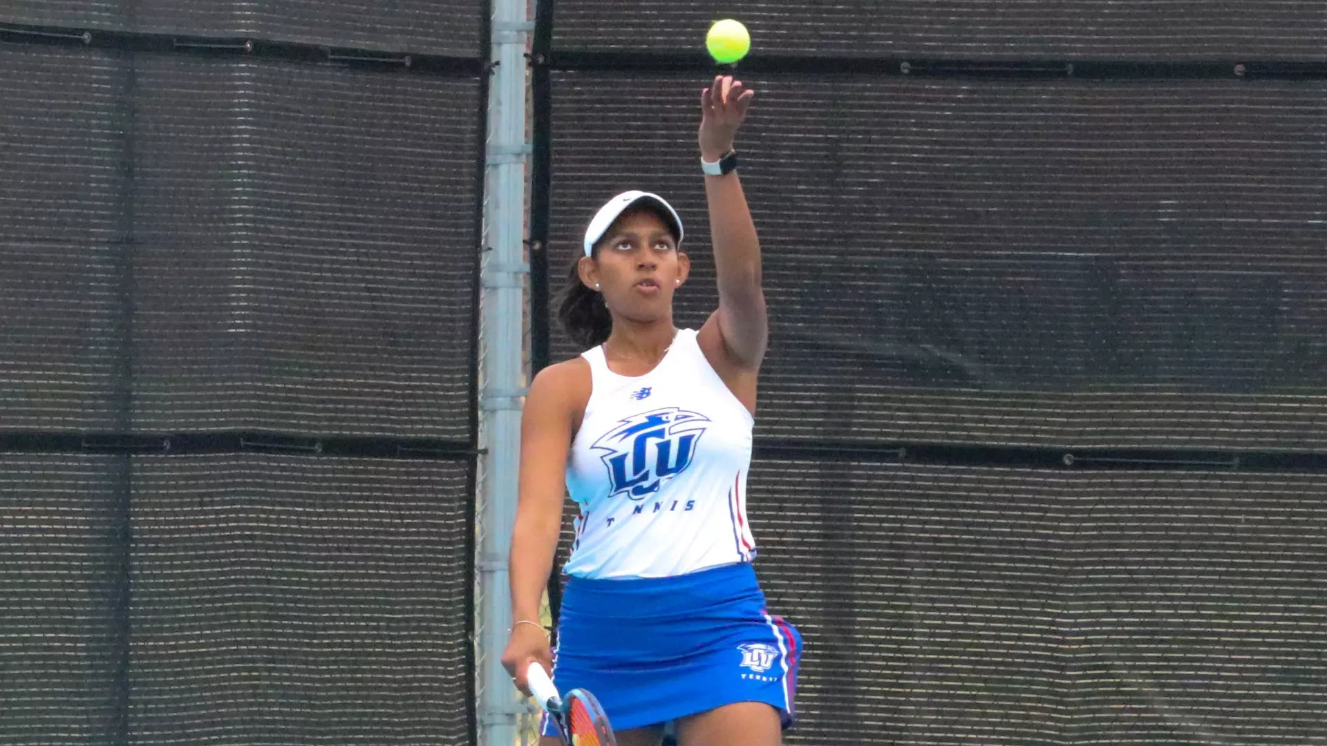 A female tennis player preparing to serve, captured mid-toss with a tennis ball visible above her raised left hand. She is wearing a white sleeveless top and blue skirt with 