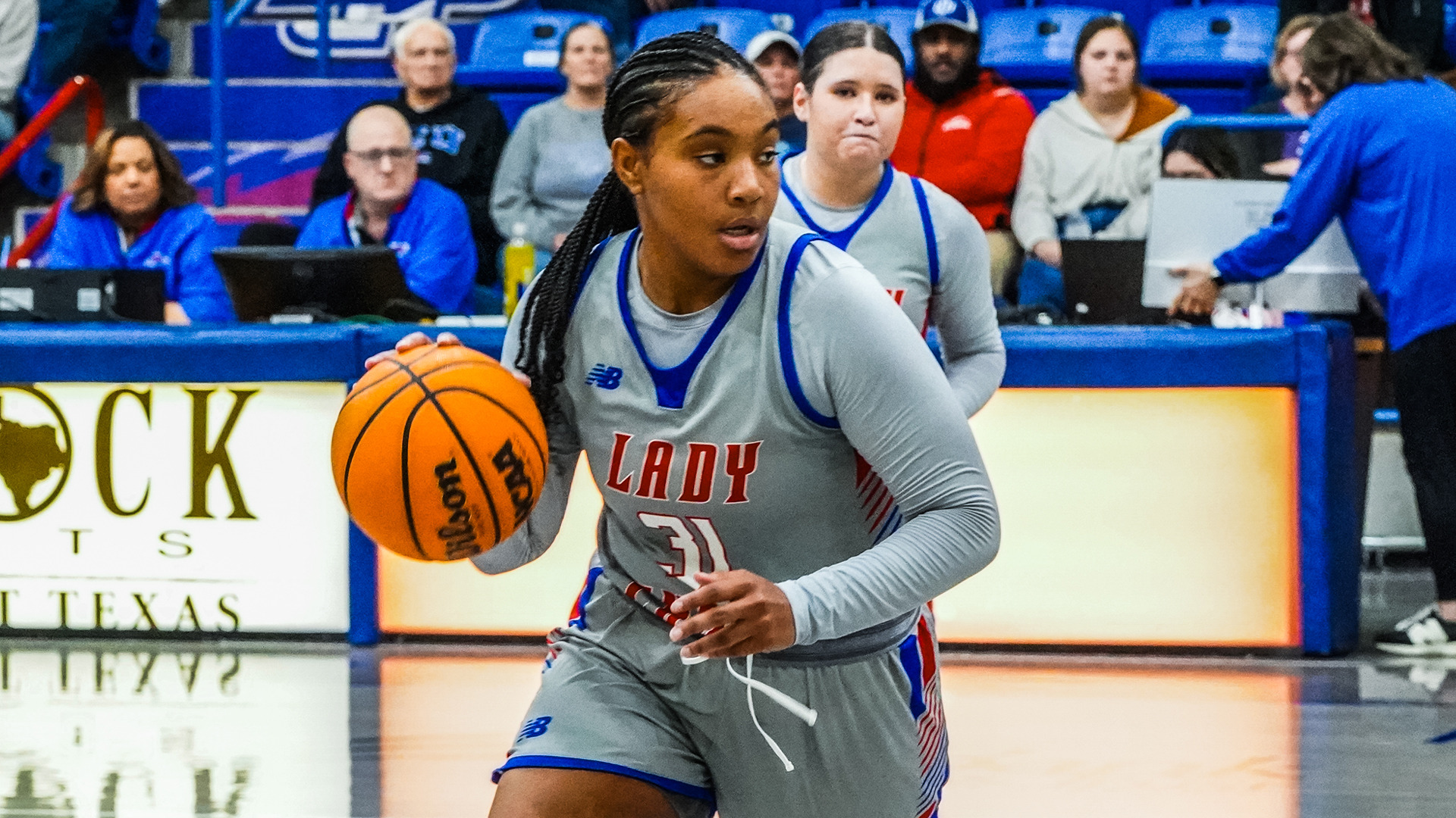 Female basketball player in gray Lady Chaps uniform #31 with blue and red trim dribbling Wilson basketball, dark braided hair pulled back. Gray long sleeve compression shirt under jersey, focused expression looking forward. Teammate in matching gray uniform visible behind her. Blue bleachers with spectators in background wearing mix of blue, gray, and red clothing. Yellow digital signage visible courtside. Game action shot showing player bringing ball up court.