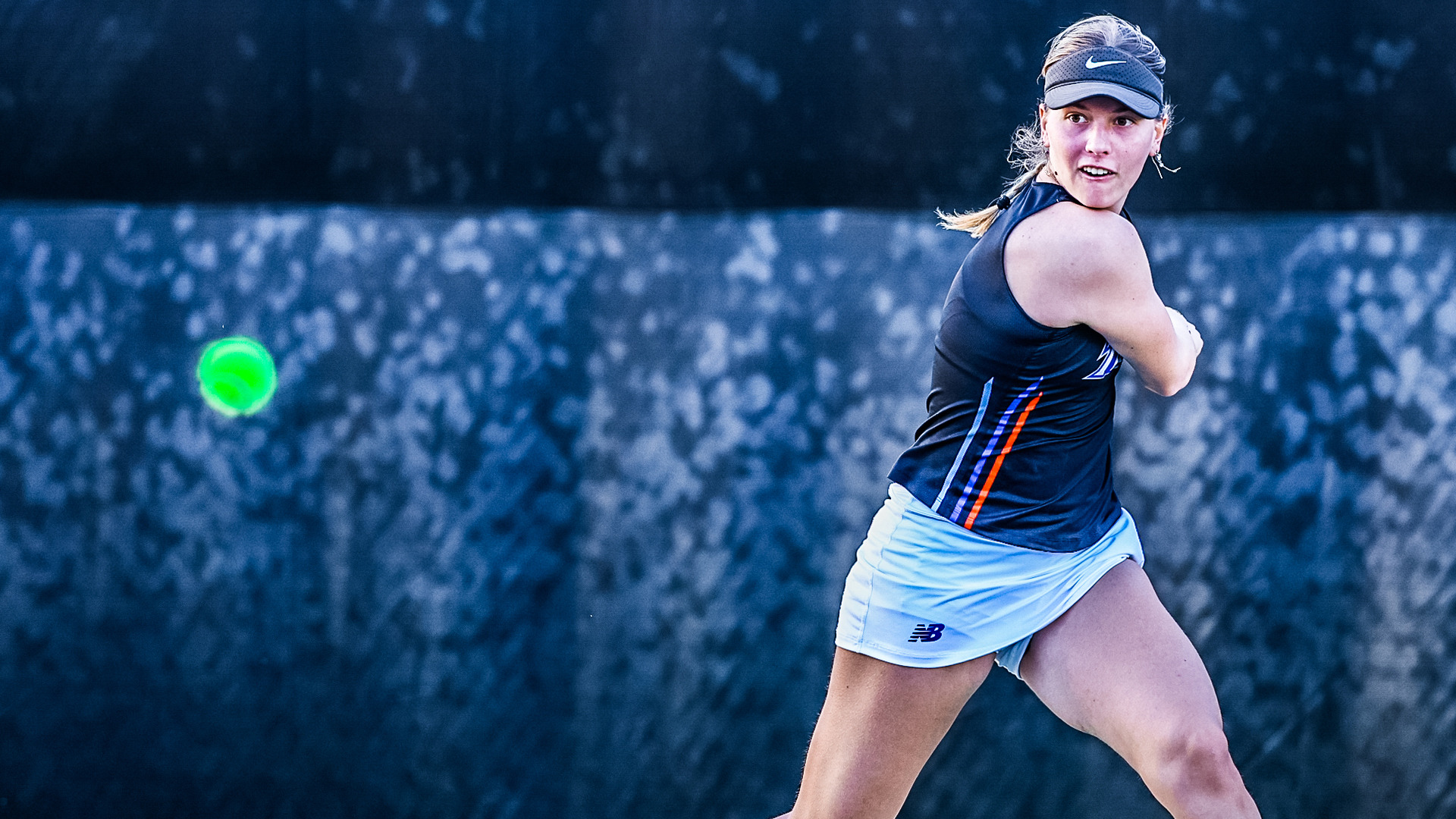 Female tennis player in dark navy sleeveless top with blue and orange accents, light blue New Balance skirt, and gray Nike visor with ponytail. Focused expression mid-swing watching bright green tennis ball in flight to her left. Dark blurred background with bokeh effect suggesting outdoor court setting with foliage. Action shot capturing powerful follow-through motion during match play.