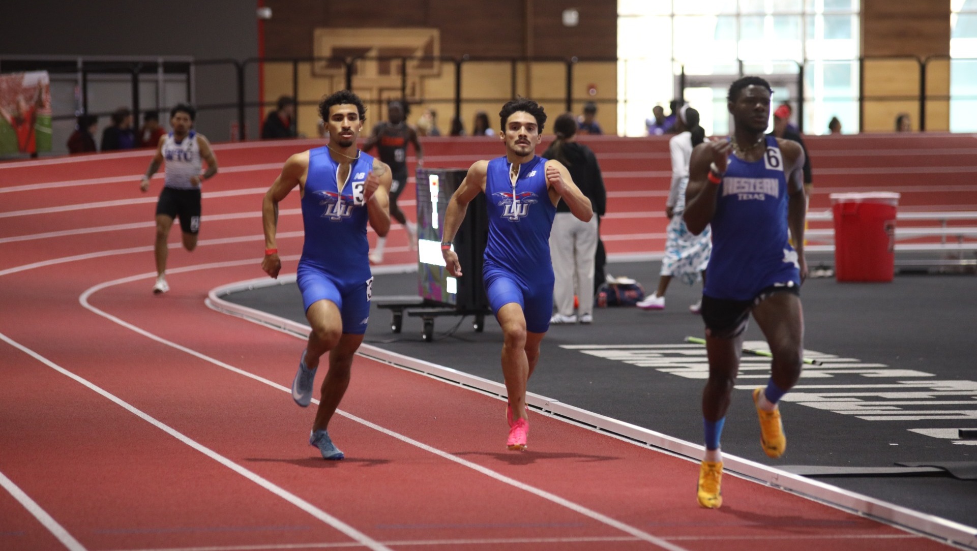 Two Lubbock Christian runners (blue uniforms) run hard to catch an athlete from Western Texas College on a red track at the Sport Performance Center at Texas Tech. 