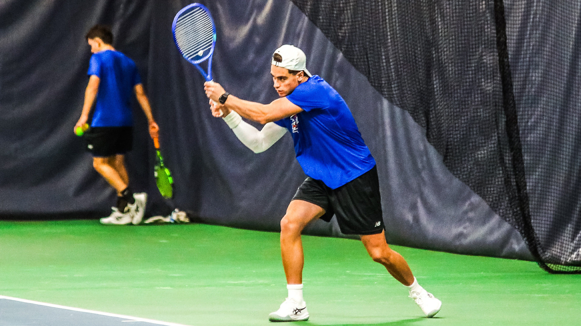 Male tennis player in blue shirt and black New Balance shorts preparing backhand shot with blue tennis racket, white cap, white compression arm sleeve on left arm. White socks and shoes. Green indoor court surface. Teammate in blue shirt visible in background left holding tennis ball with green racket. Dark curtain backdrop behind players.