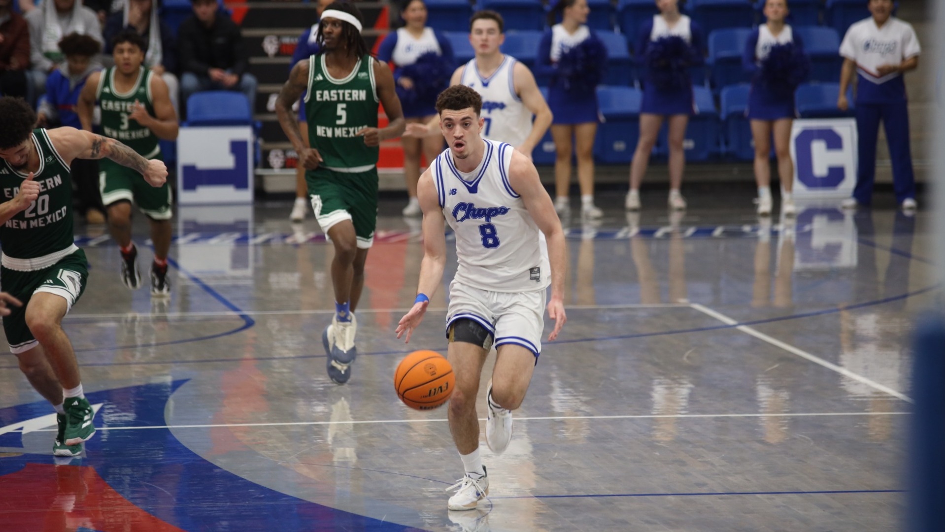 This is a photo of Lubbock Christian forward Amondo Miller Jr. (white jersey, #8) dribbling the basketball up the court on a fastbreak in a game against Eastern New Mexico (green jerseys) at the Rip Griffin Center. 