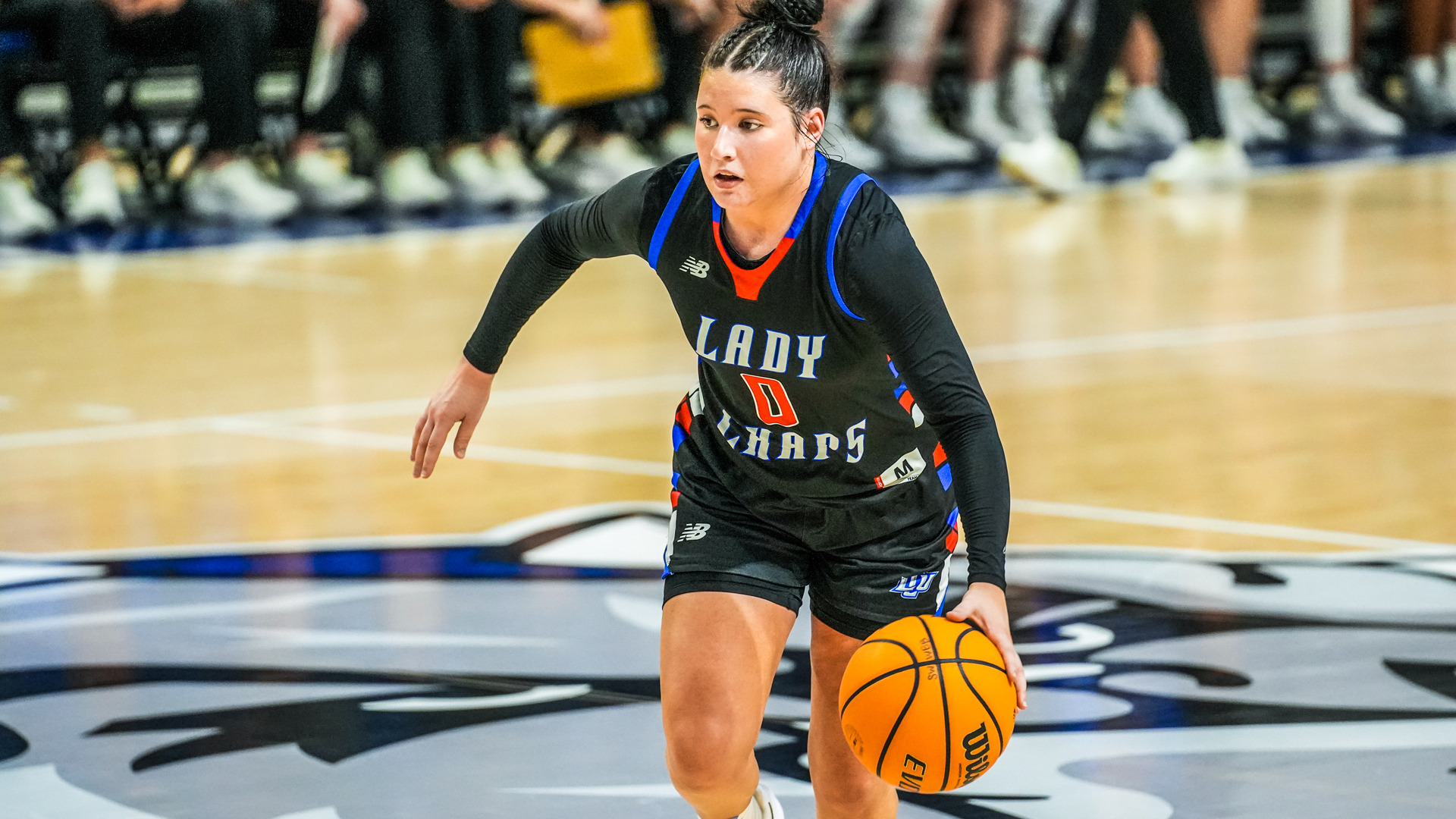 Female basketball player in black Lady Chaps uniform #0 with blue and orange trim dribbling Wilson basketball on light-colored court. Dark hair pulled back in high bun, focused expression, athletic stance with left arm extended for balance. New Balance logos visible on jersey and shorts. Blurred background shows spectators and officials along sideline, yellow signage visible in distance. Action shot capturing player bringing ball up court.