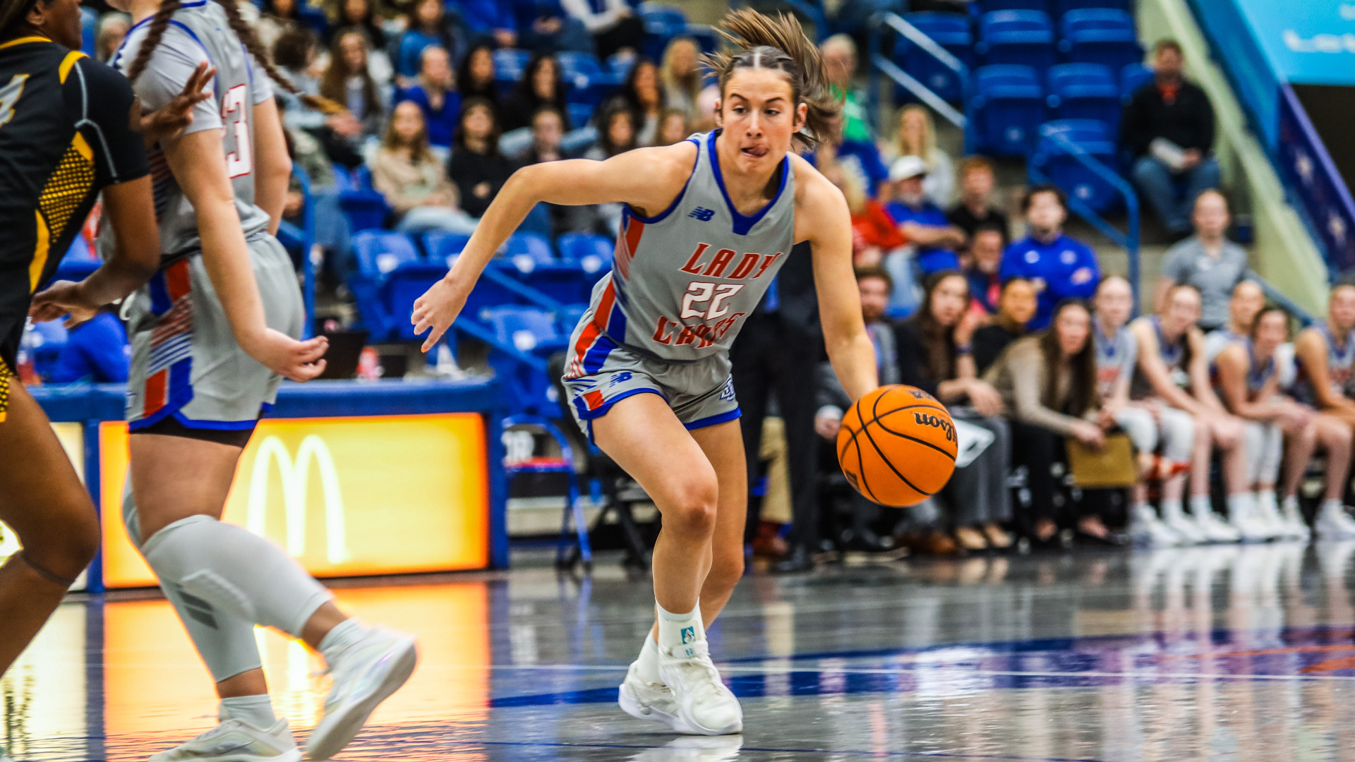 LCU women's basketball player number 22 in white 'LADY CHAPS' uniform with New Balance logo dribbling Wilson basketball downcourt at high speed. Hair flying back showing motion. Blue arena seating filled with spectators in background. McDonald's golden arches advertising board visible courtside on left. Opponents in black and yellow uniforms defending. Glossy hardwood court reflects arena lights