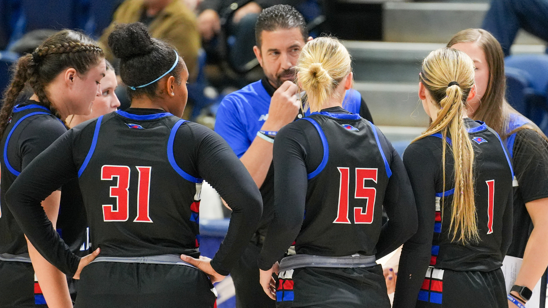 Male coach in blue shirt speaking to huddle of Lady Chaps basketball players in black uniforms with blue and red trim, viewed from behind. Visible jersey numbers include 31, 15, and 1. Players with various hairstyles including braids, bun, and ponytails listening to coach's instructions during timeout. Gray bleachers and spectators visible in blurred background. Team meeting during game with focused attention on coach's guidance.