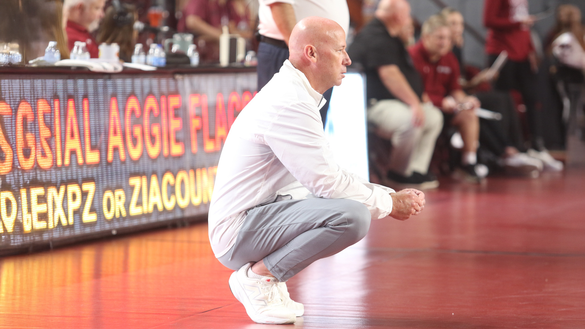 Bald male coach in white dress shirt and gray pants crouching on sideline during basketball game, hands clasped, intently watching play. Digital courtside display with orange text partially visible behind him showing social media information. Water bottles on scorer's table in background. Blurred crowd and opposing team members visible in background. Coach in focused, attentive position observing game action on polished wooden court.
