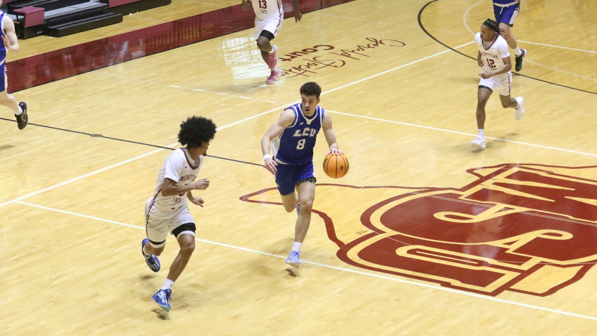 This is a photo of Lubbock Christian men's basketball forward Amondo Miller Jr. (blue jersey, #8) dribbling the ball on the fastbreak toward a Midwestern State defender (white jersey) at D.L. Ligon Coliseum.