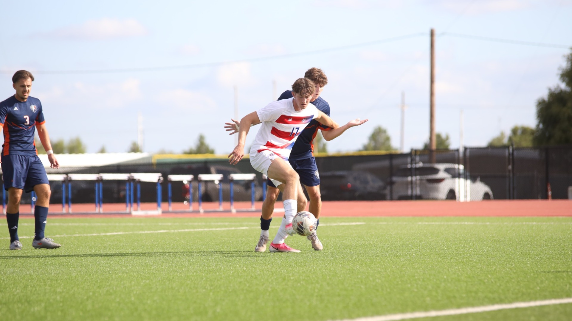 This is a photo of Lubbock Christian forward Jack Brassey (white jersey, #17) holding off a UT Tyler defender (navy blue jersey) while receiving a soccer pass.