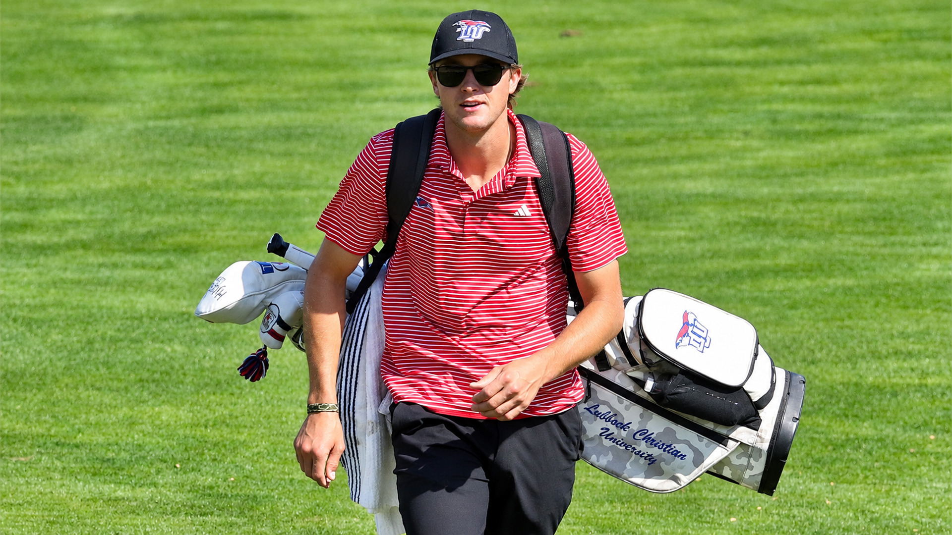 Golfer in a black LCU cap, dark sunglasses, red-and-white striped polo, and black pants walks down a sunlit fairway, carrying a camouflage golf bag that reads 