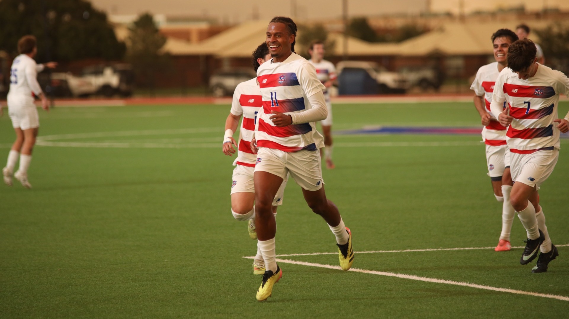 This is a photo of Lubbock Christian forward Augusto Militao (white jersey, #11) smiling and running across the field after scoring a goal with teammates following behind him.