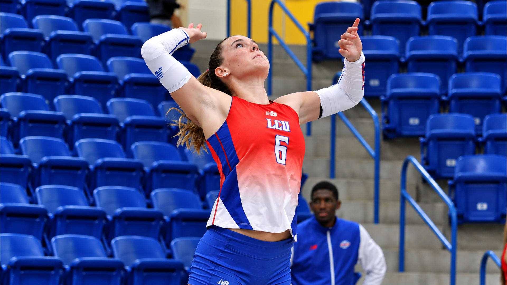 Action photo of LCU player #6 in a red-to-white jersey with blue trim and white arm sleeves. She is mid-serve, with both arms raised overhead and her eyes on the ball above her. A staff member in a blue and white jacket watches from the right. Rows of empty blue stadium seats and concrete steps with blue railings fill the background.