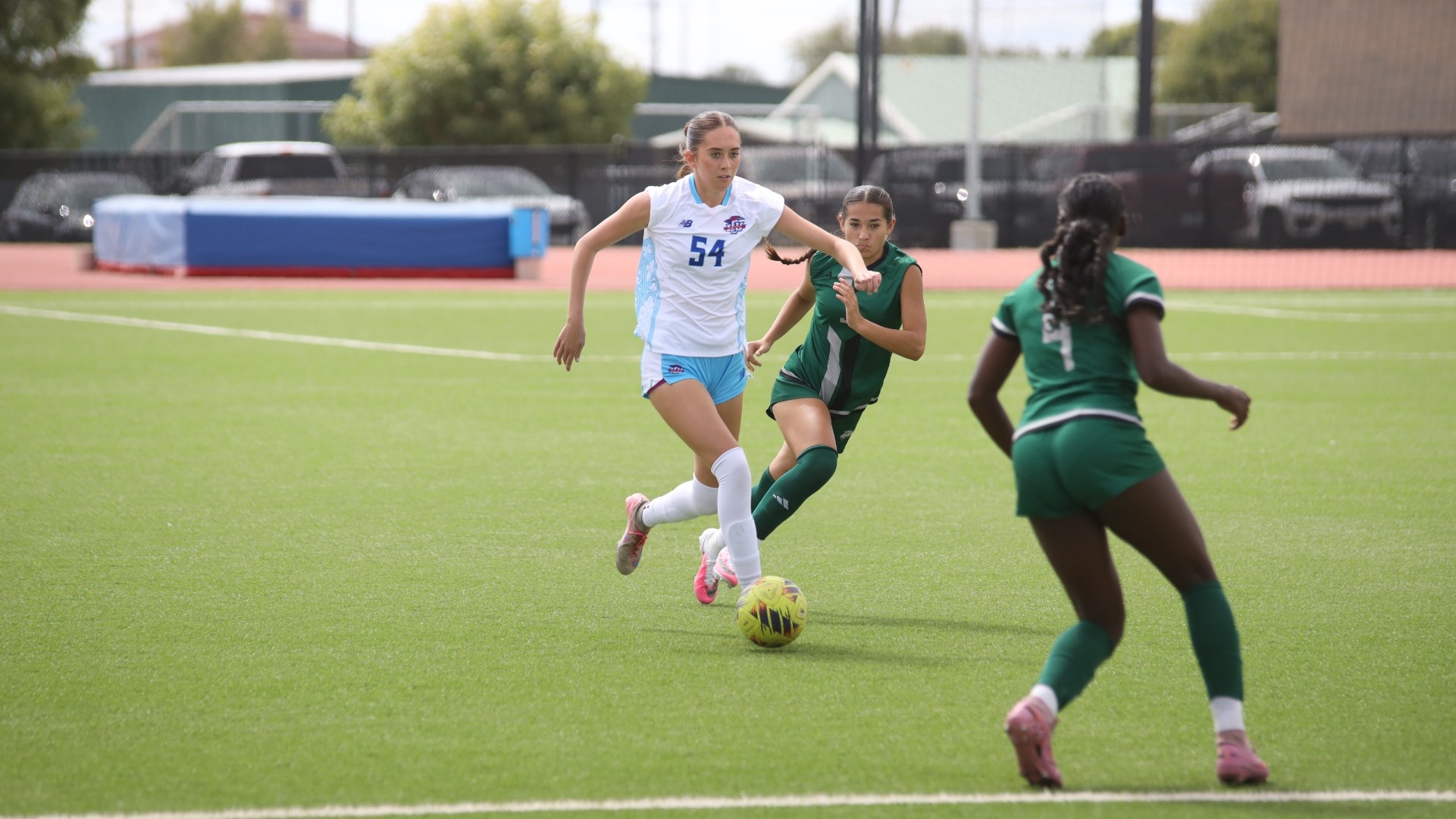 This is a photo of Lubbock Christian defender Araceli Patterson (white jersey, #54) dribbling a yellow soccer ball in between a pair of Eastern New Mexico players (green jerseys).