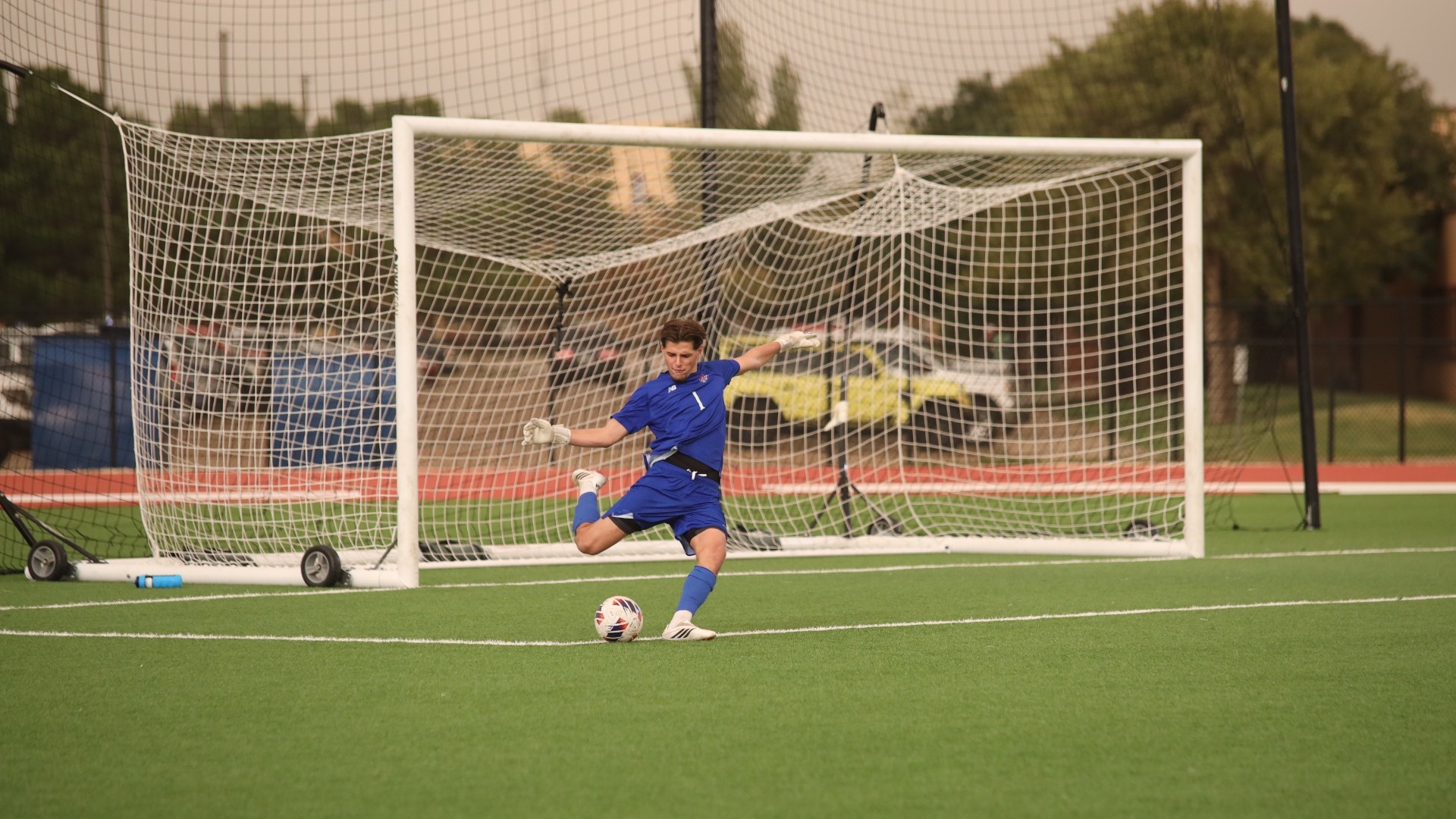 This is a photo of Lubbock Christian goalkeeper Milo Paci (blue jersey, #1) preparing to kick a soccer ball in front of the goal on a green turf field.