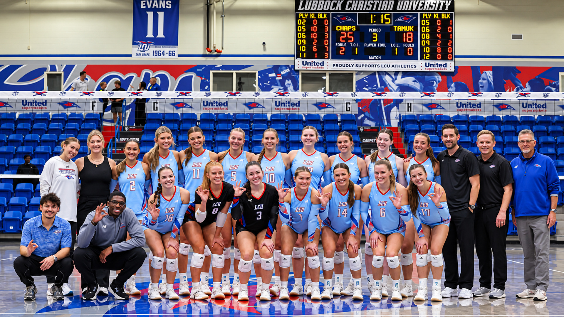 Group photo of the LCU volleyball team and staff posed in rows on the court, most smiling and many making an “OK” hand gesture. Players wear light blue jerseys, with two in black jerseys numbered 8 and 3, and others numbered 2, 11, 5, 10, 14, 6, and 17. Staff members in team apparel stand and crouch at both ends of the group. Behind them are blue stadium seats and a large mural. A scoreboard overhead reads “Lubbock Christian University,” with the Chaps leading 25 to 18 in period 3, and a retired jersey banner reading “Evans 11” hangs at left.