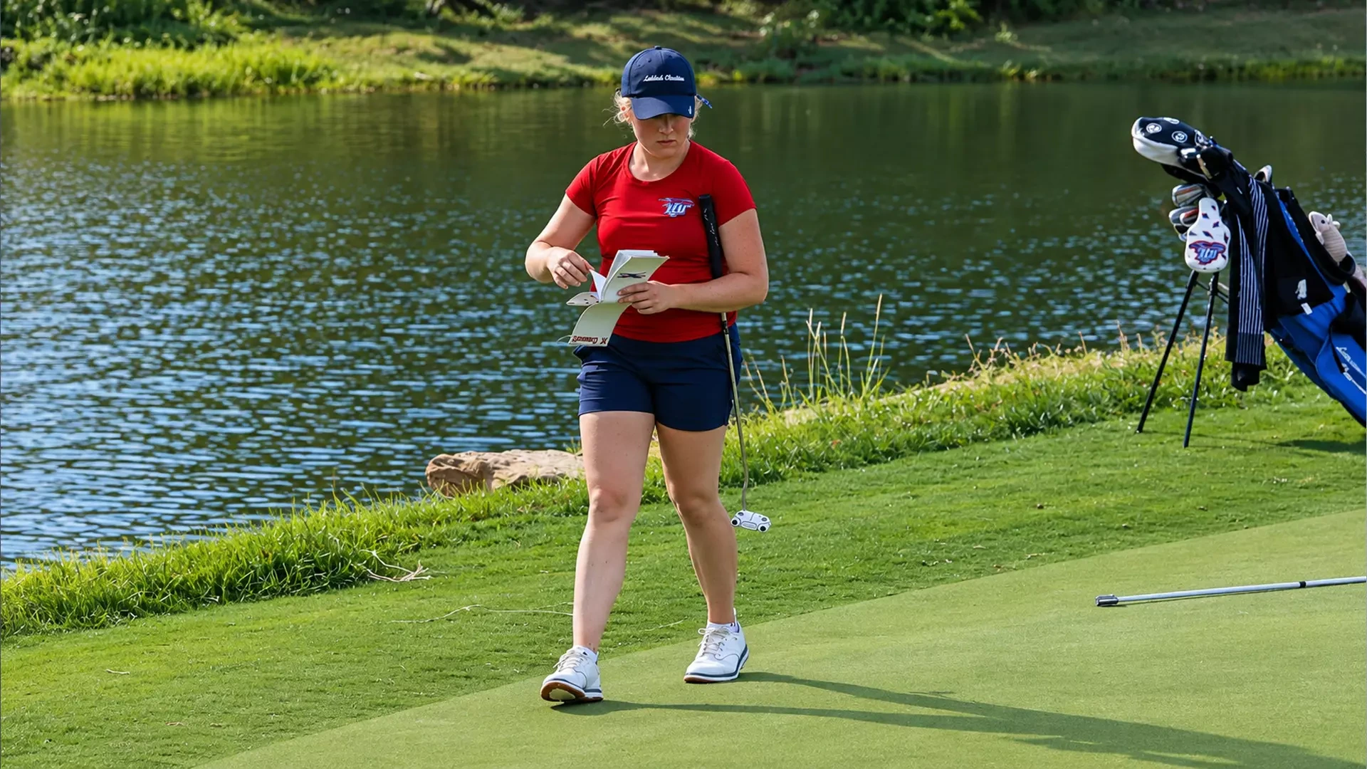 Golfer in a navy cap, red shirt, and navy shorts walks across a green beside a pond, looking down at a notebook in her hands with a putter tucked under her arm. A golf bag stands on the right, and a club lies on the grass nearby. Water and green grass fill the background.