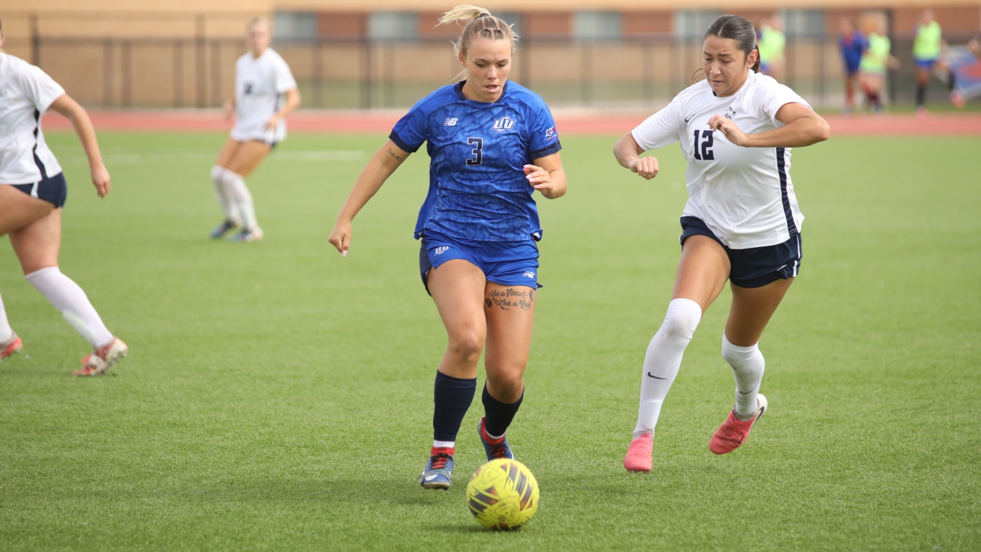 This is a photo of Lubbock Christian forward Logan Holloway (blue jersey, #3) dribbling a yellow soccer ball with a St. Edward's defender (white jersey, #12) chasing her.