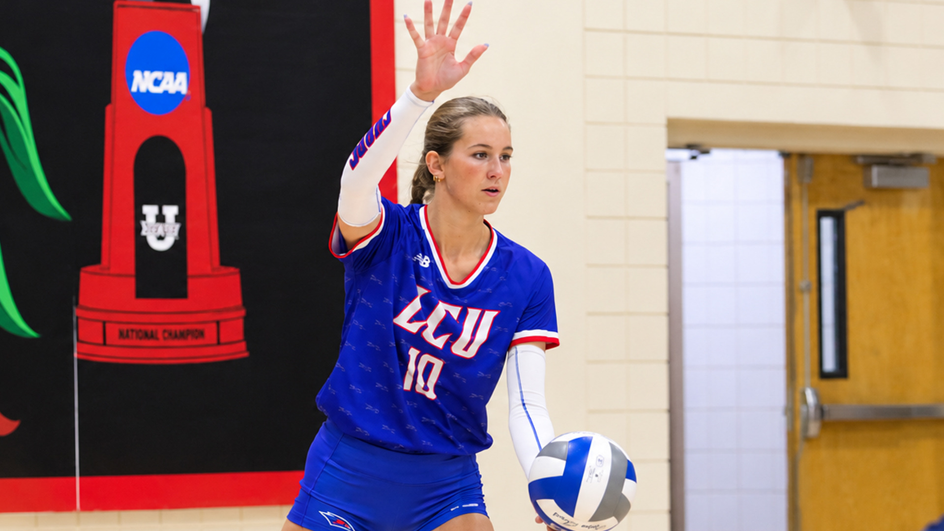 Action photo of LCU player #10, in a royal blue jersey with red and white trim and white arm sleeves, preparing to serve. She holds a volleyball low in one hand and raises the other hand, palm open, with a focused expression. Behind her, a red and black 
