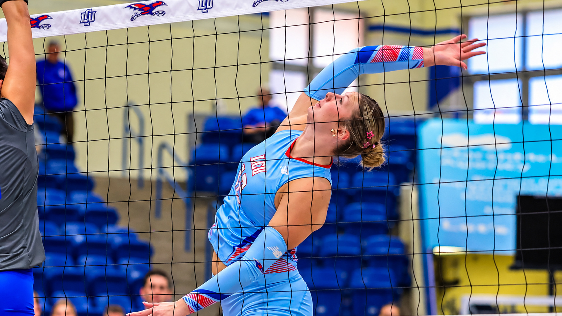 Action photo taken through the net during a volleyball match. An LCU player in a light blue jersey arches back with one arm extended behind her, a pink star clip in her hair, as she swings to attack the ball. A hand and arm from an opposing player reach up at the far left. Blue stadium seating, spectators, and a large banner fill the background.
