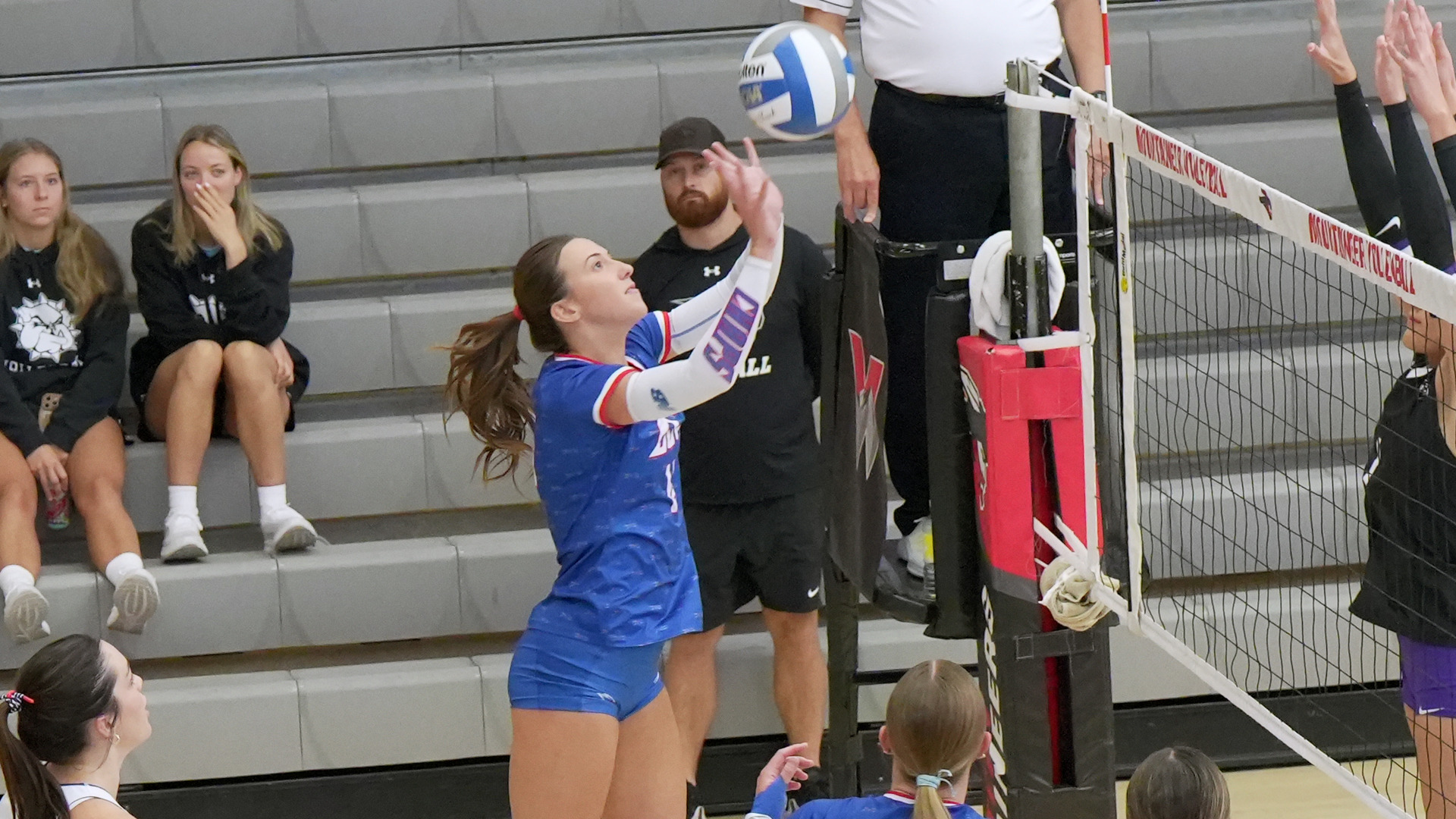 Action photo taken courtside near the net. An LCU player in a blue jersey reaches up to set the ball, with an opposing player blocking from the other side of the net marked 