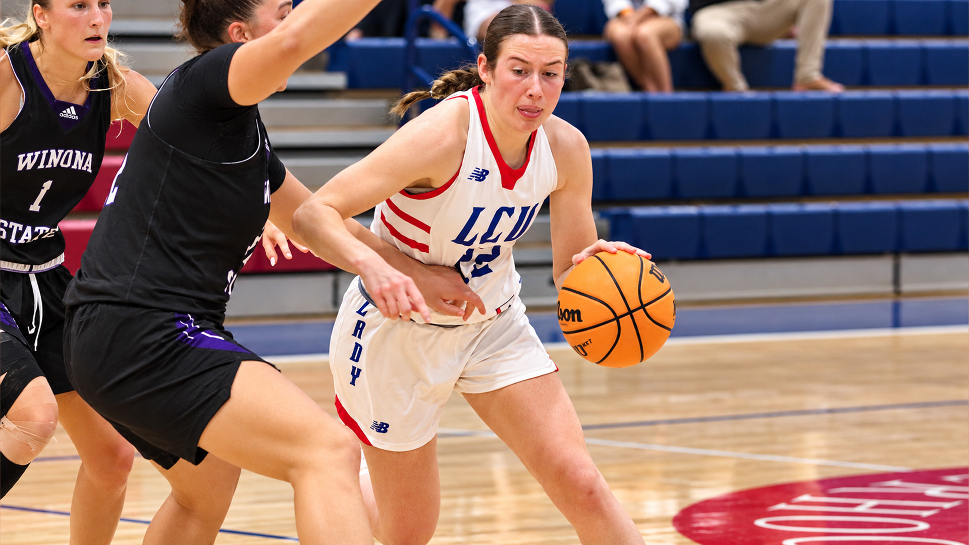 Action photo of an LCU basketball player in a white “Lady” uniform, number 12, driving toward the basket while dribbling with her right hand. Two defenders in black Winona State jerseys close in around her, one reaching an arm across her body. Blue bleachers and a few spectators are visible in the background, and a red logo is painted on the court at lower right.
