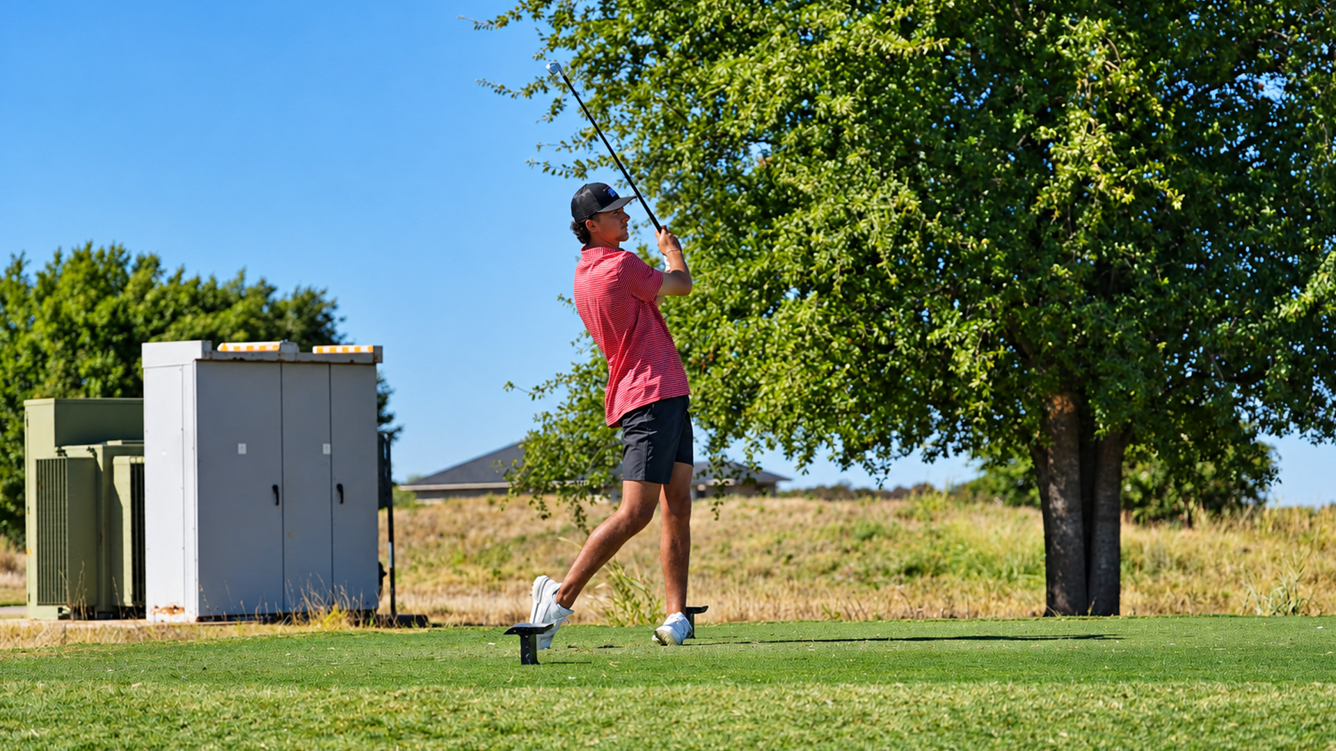 Golfer in a red striped polo and dark shorts completes his swing on a tee box, watching the ball's flight. A large tree and open grassy field with a house in the distance fill the background, under a clear blue sky.