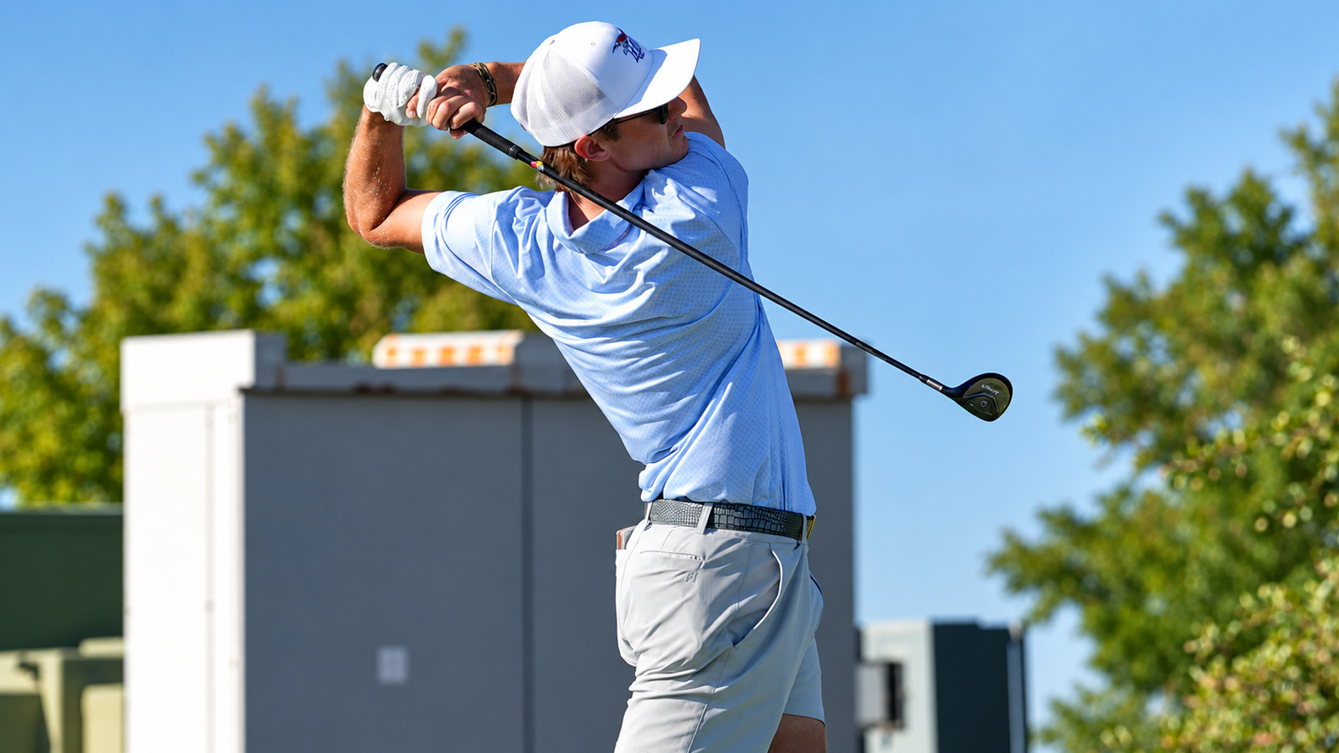 Golfer in a light blue polo and gray shorts completing his swing follow-through, looking upward to track the ball's flight. Trees and a utility structure are visible in the background under a clear blue sky.